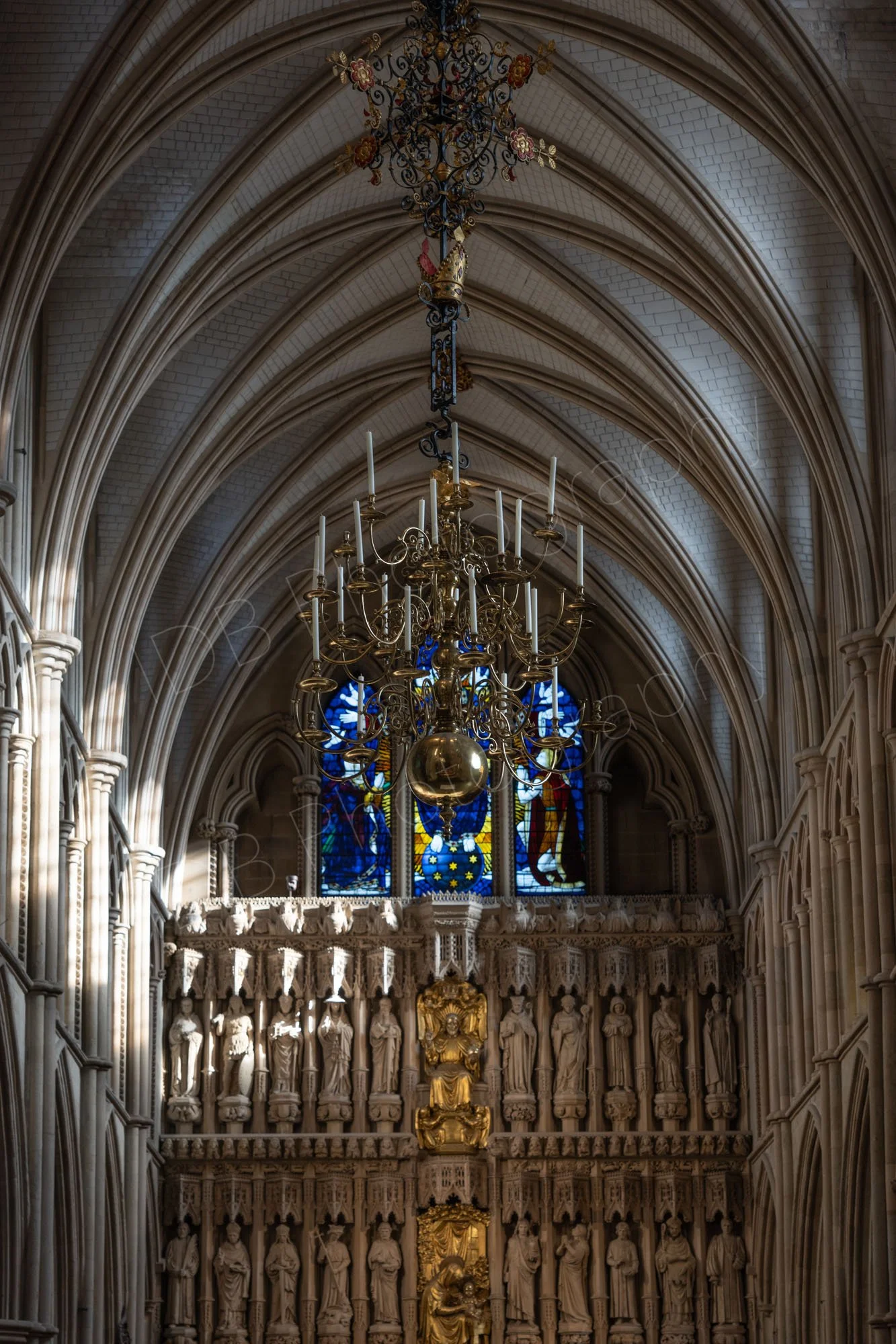 A beautiful vaulted ceiling of Southwalk Cathedral. A number of carved stone figures line the wall; a spectacular chandelier and stained glass window can be seen.