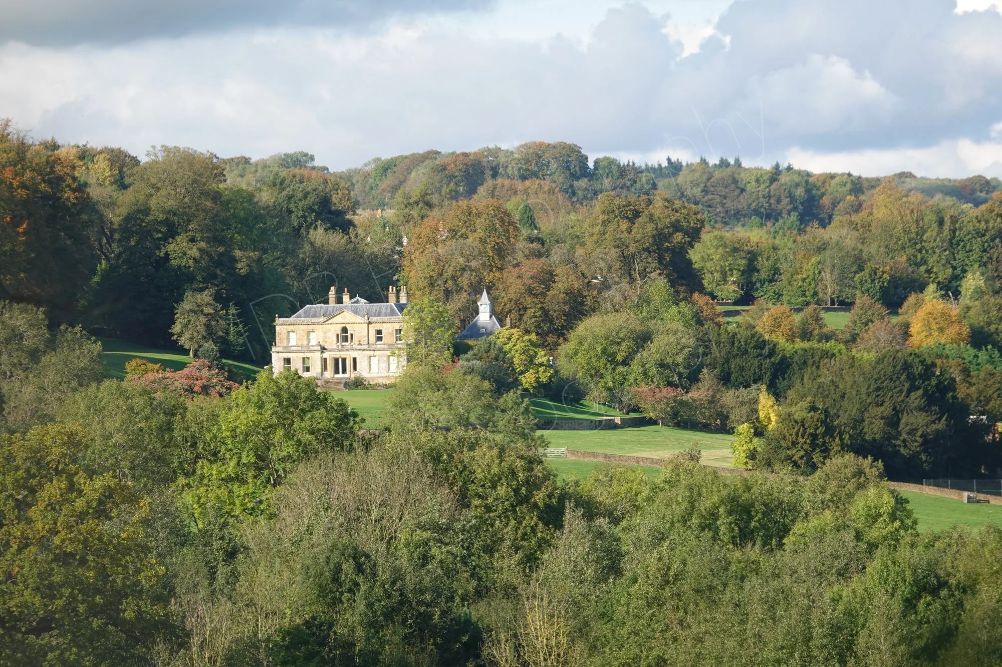 A beautiful view of the Cotswold countryside with a large Cotswold stone manor house and gardens set amongst attractive woodland under a cloudy shy.