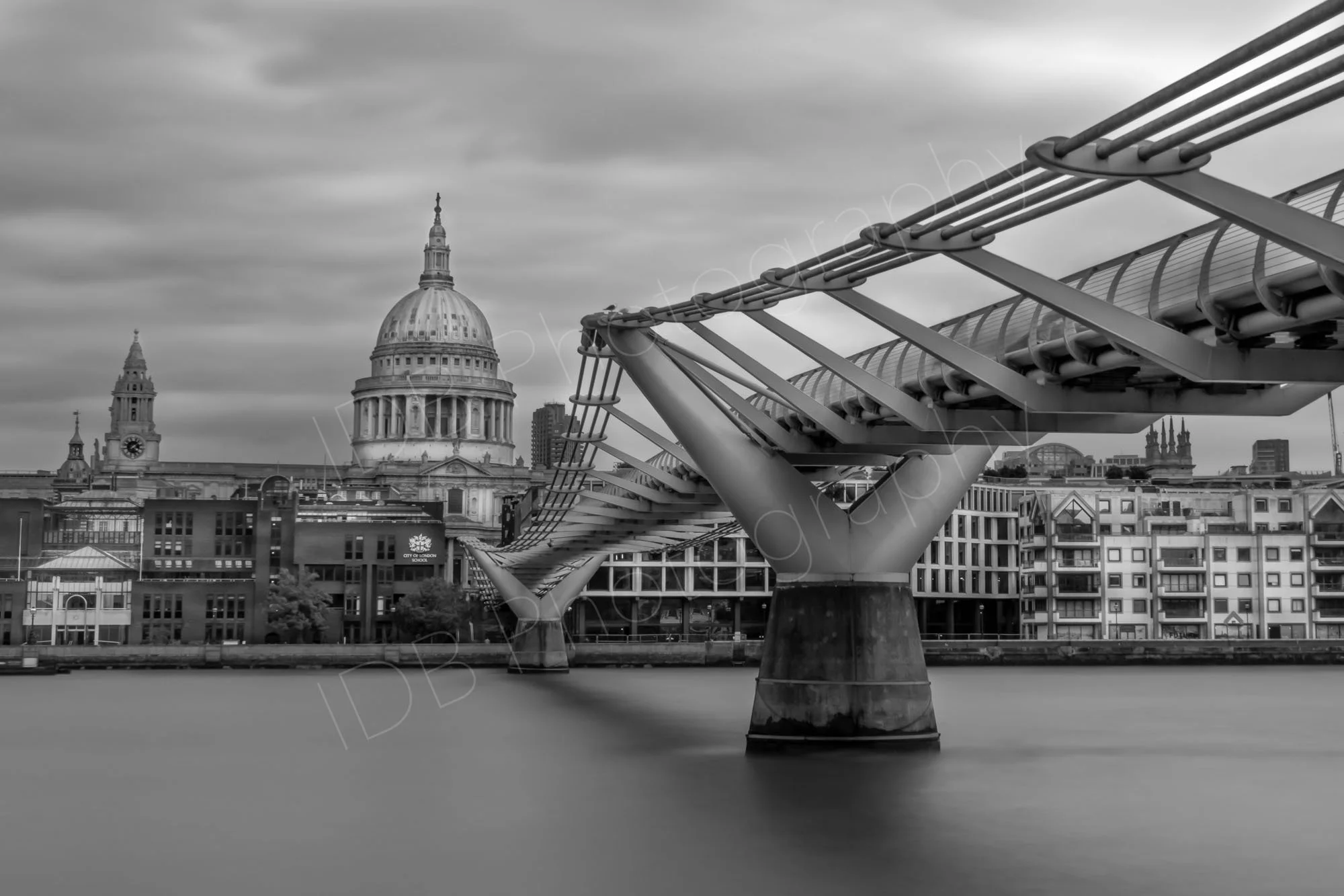 A black and white version of a long ND filter exposure across the Thames with the Millennium Bridge and St Paul's Cathedral clearly seen; smooth water and cloudy sky.
