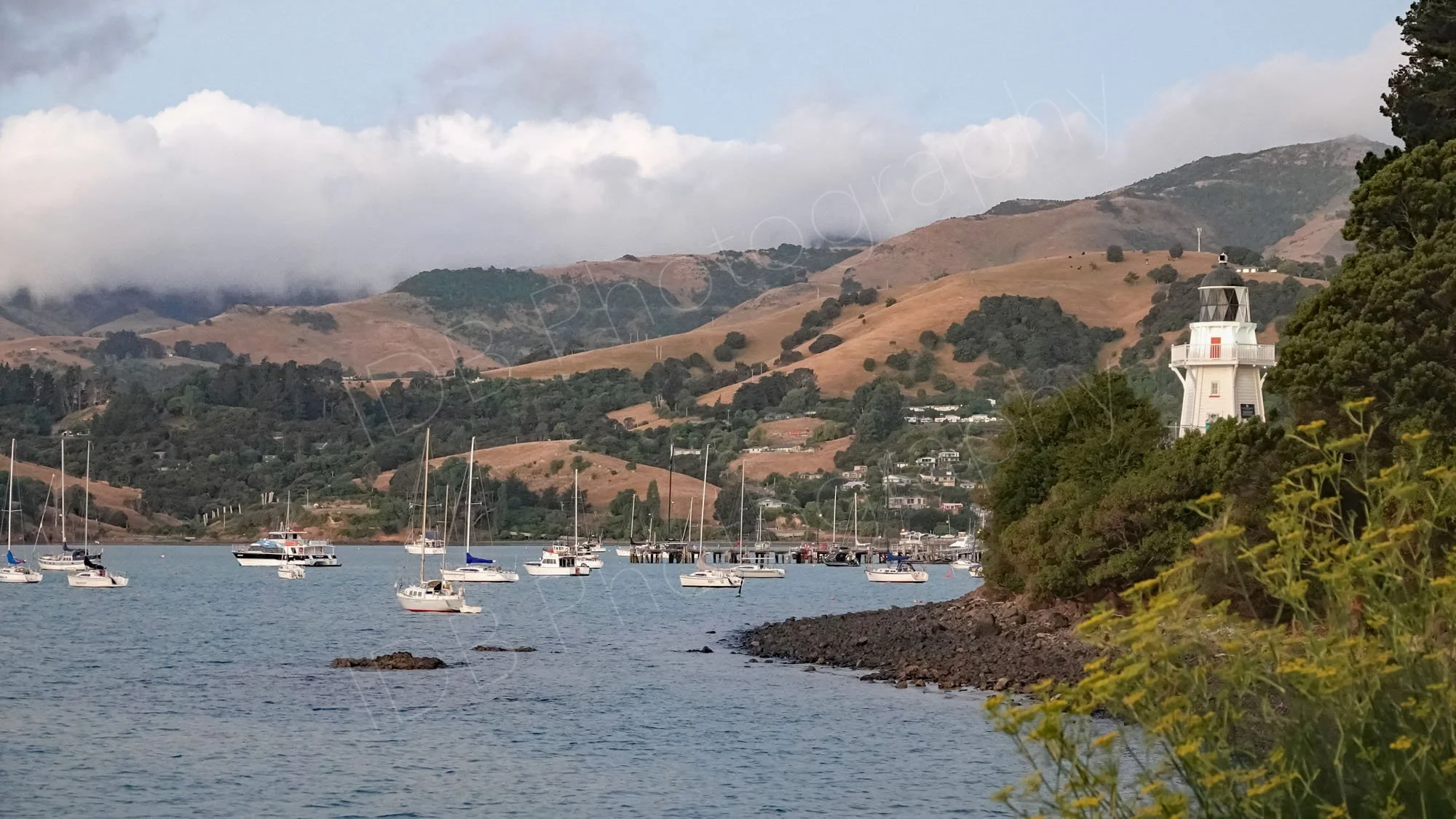 Akaroa Harbour and lighthouse  with boats and a hilly cloudy background.