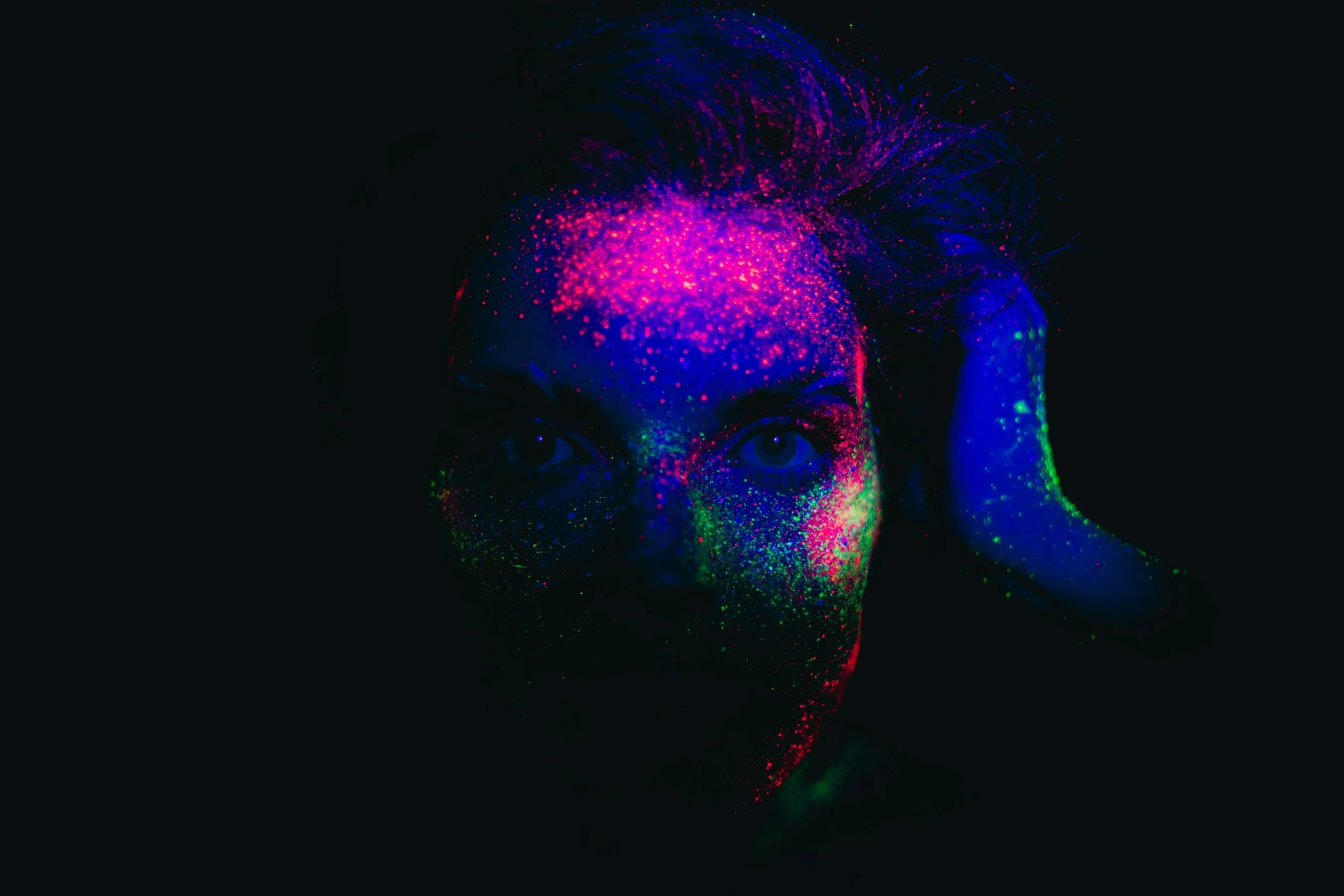 Close-up of a person's face with neon paint splattered across it, with vibrant colors including pink, blue, green, and red, on a black background.