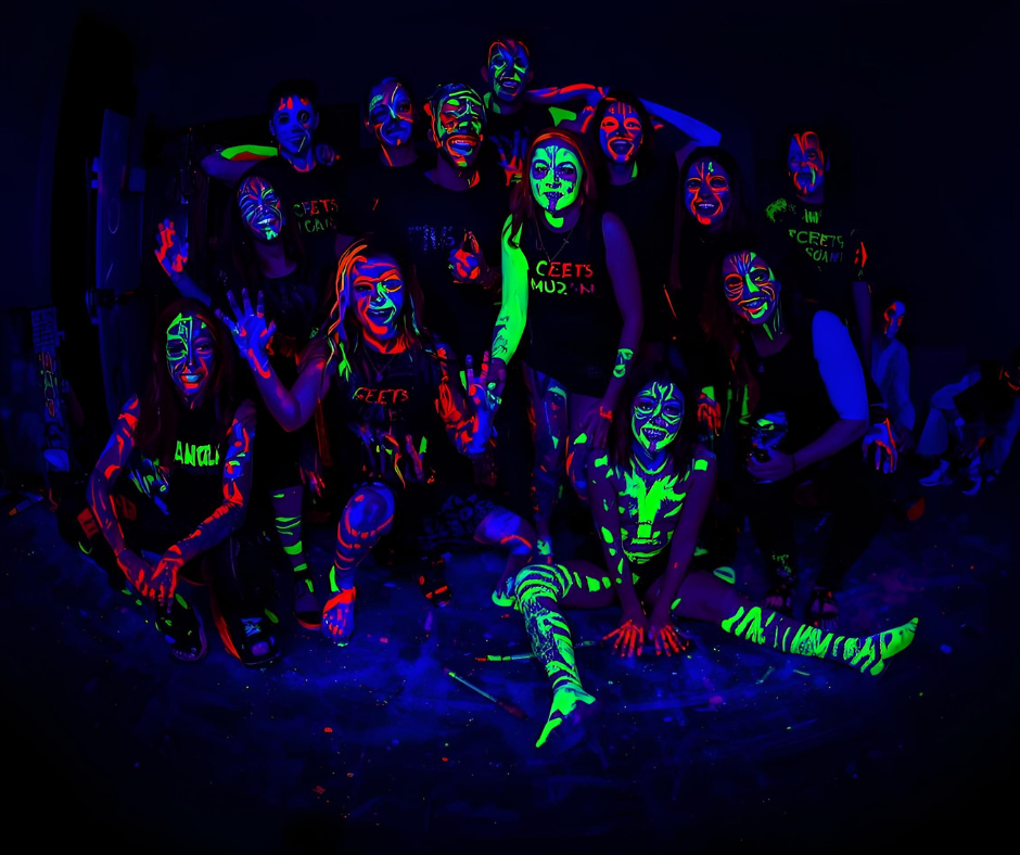 Group of people with glow-in-the-dark face and body paint under blacklight, smiling and posing.