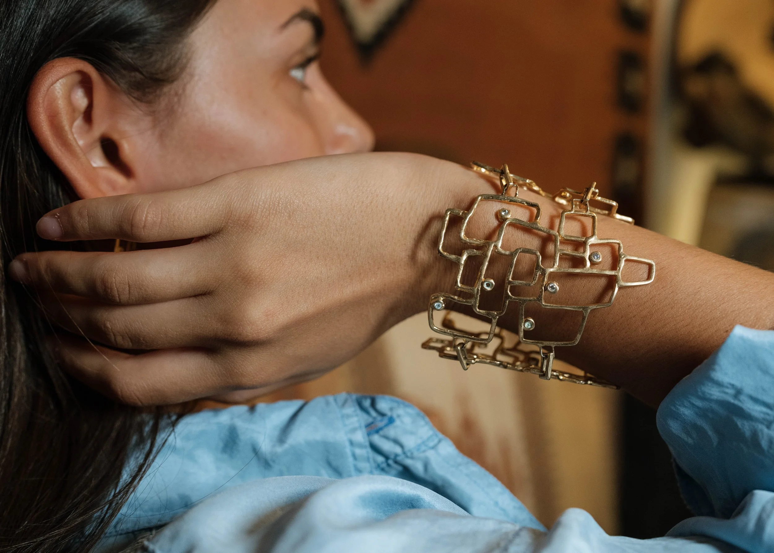 Close-up side view of a woman wearing a gold square-link bracelet with small embedded gemstones, resting her chin on her hand, with a blurred indoor background.