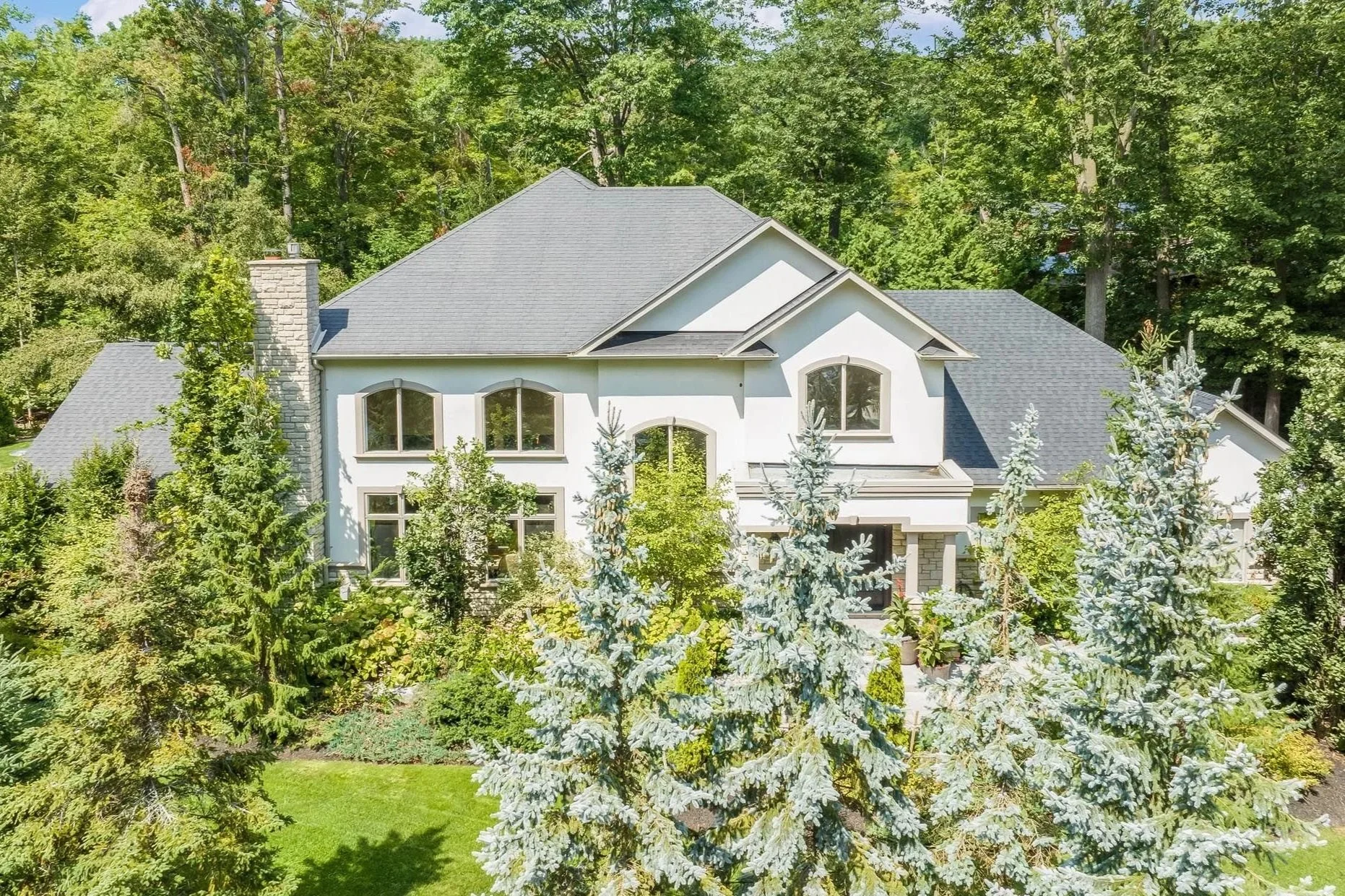 Large white house with a gray roof surrounded by lush green trees and a well-maintained lawn.