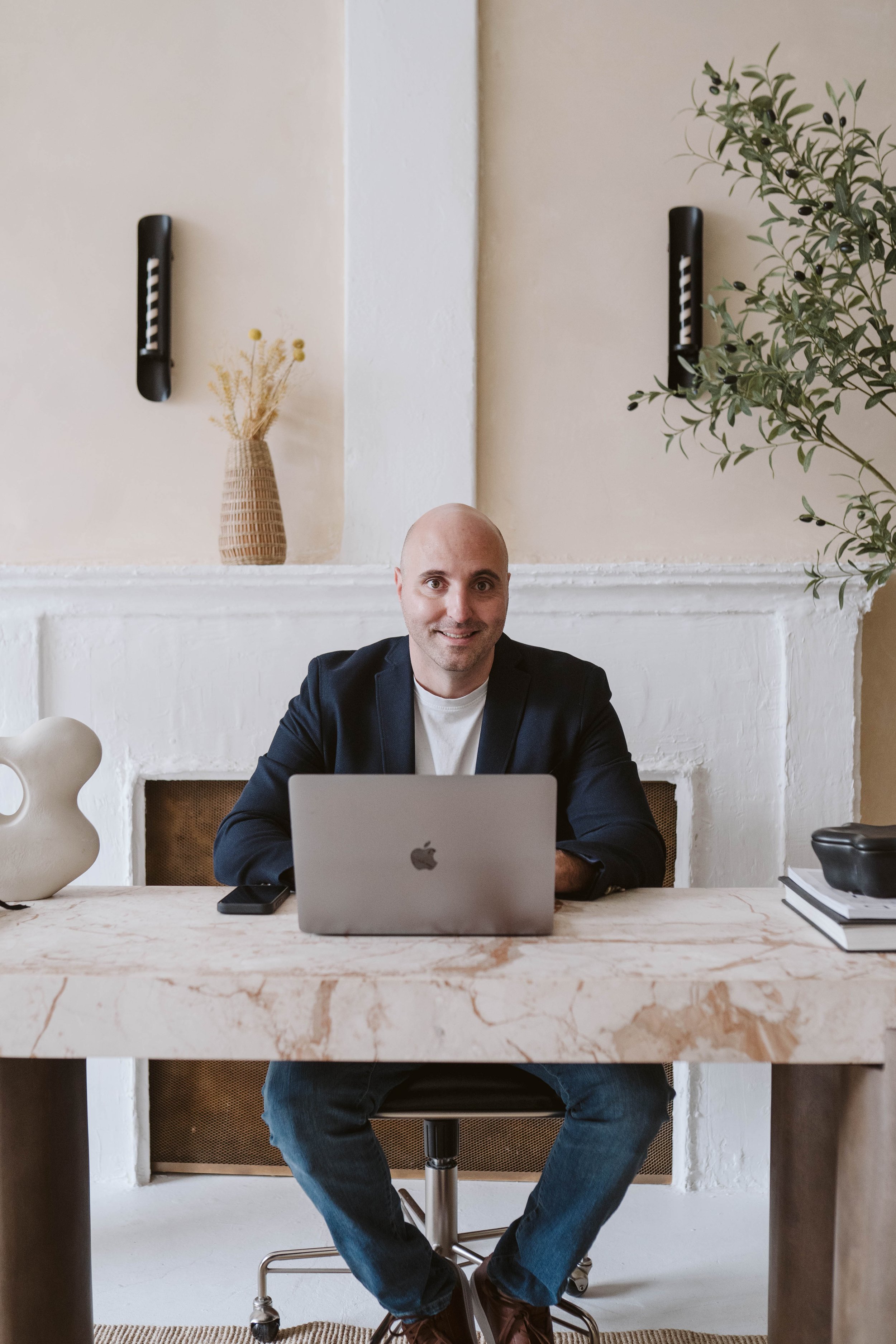 A man in a black blazer and white shirt sitting at a marble desk with a silver laptop, a smartphone, and a black bag, smiling at the camera, in a modern, minimalistic room with a large plant and decorative wall-mounted objects.