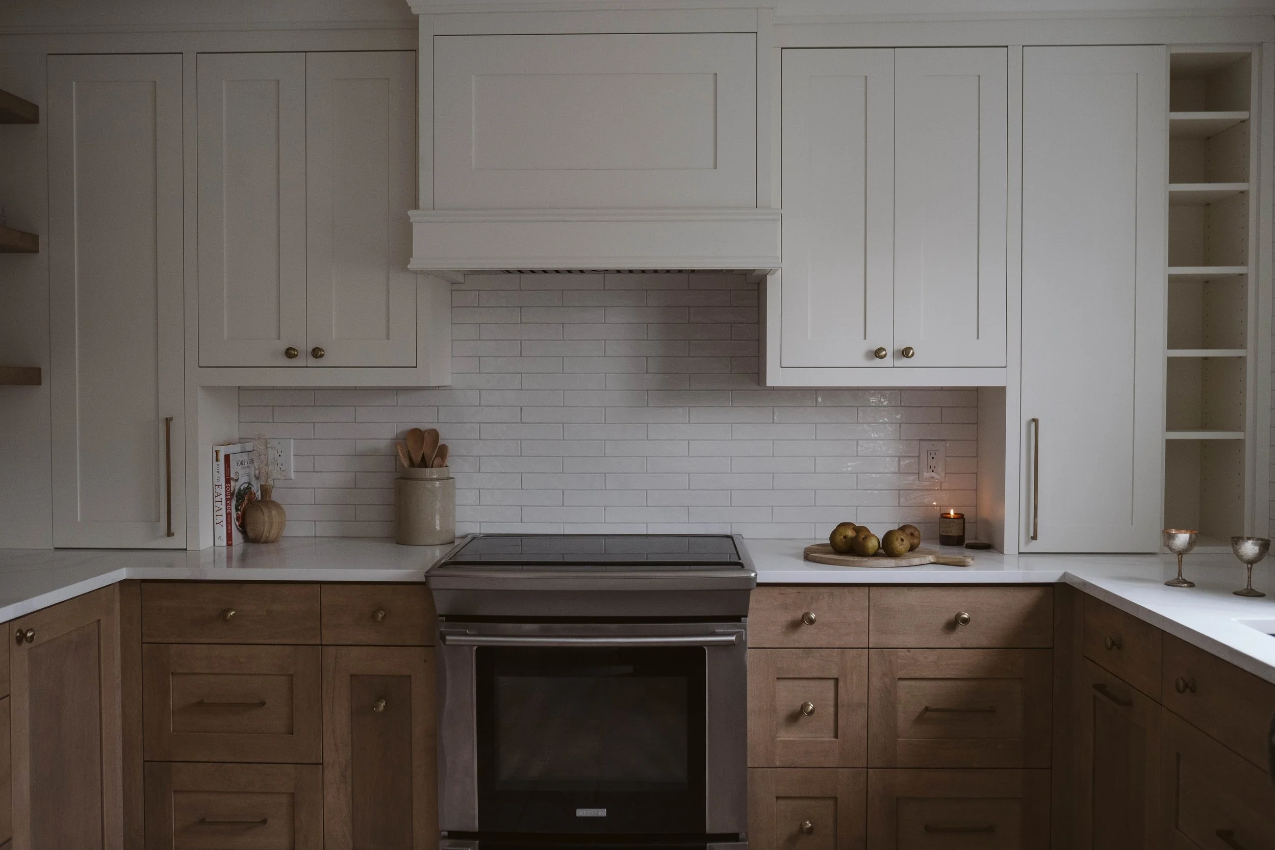 Kitchen with white upper cabinets, wooden lower cabinets, a stainless steel oven, white tile backsplash, and small decorative items including a bowl of apples, two candles, and a jar of wooden utensils.