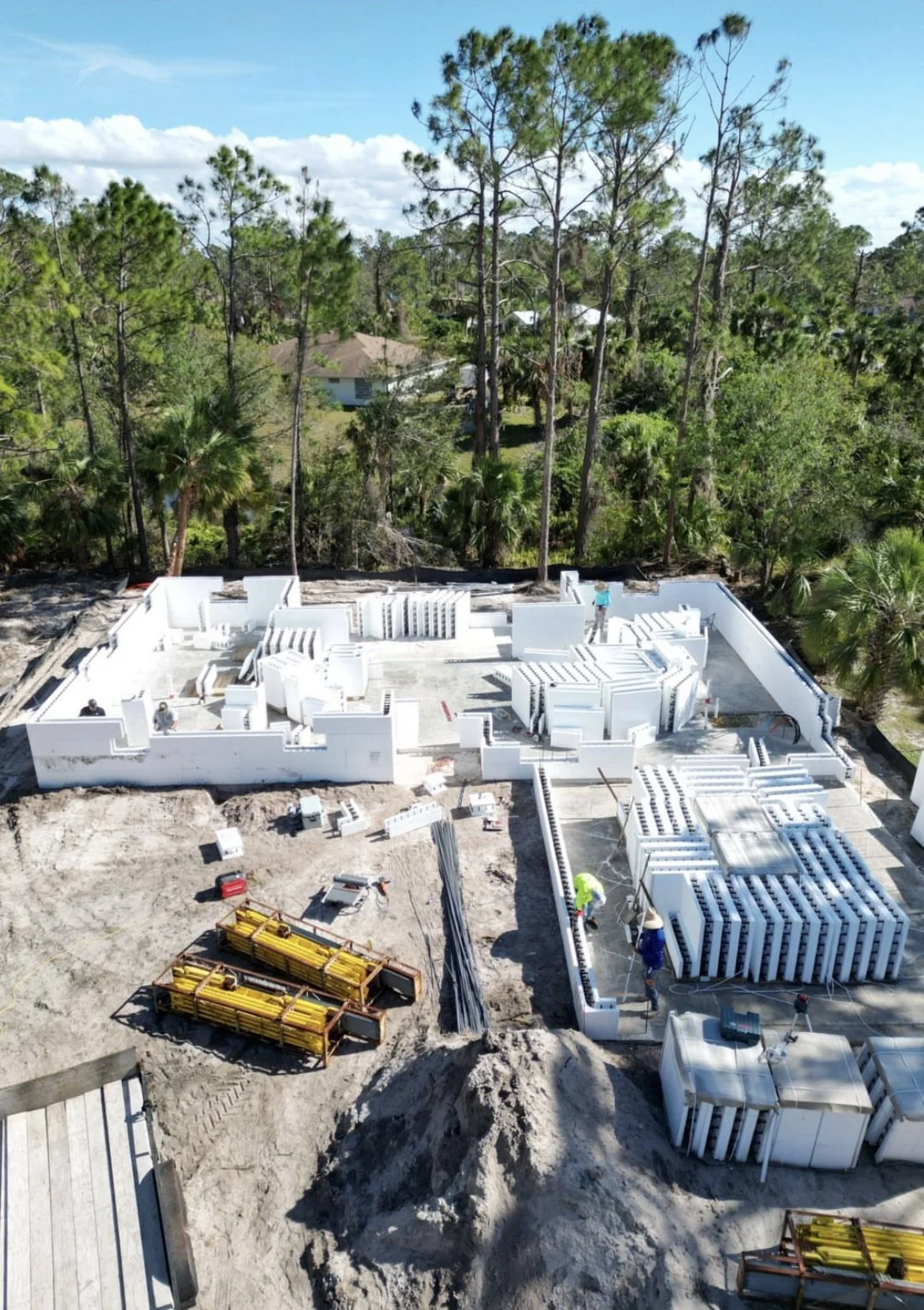 A construction site for a building in a wooded area with tall trees in the background. The foundation and walls are partially built with white materials, and construction workers are present.