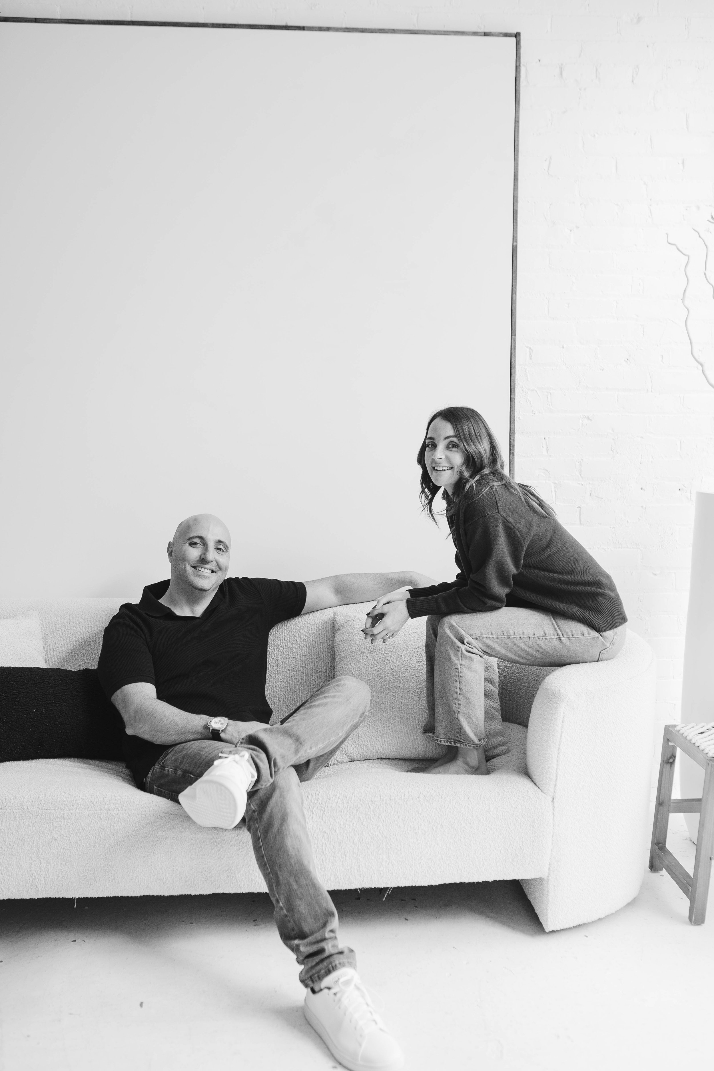 Black and white photo of a man and woman smiling, sitting on a couch in a modern, minimalistic room. The woman is sitting on the armrest of the couch, leaning towards the man, who is sitting relaxed on the sofa with his legs crossed. They are holding hands and looking at the camera.