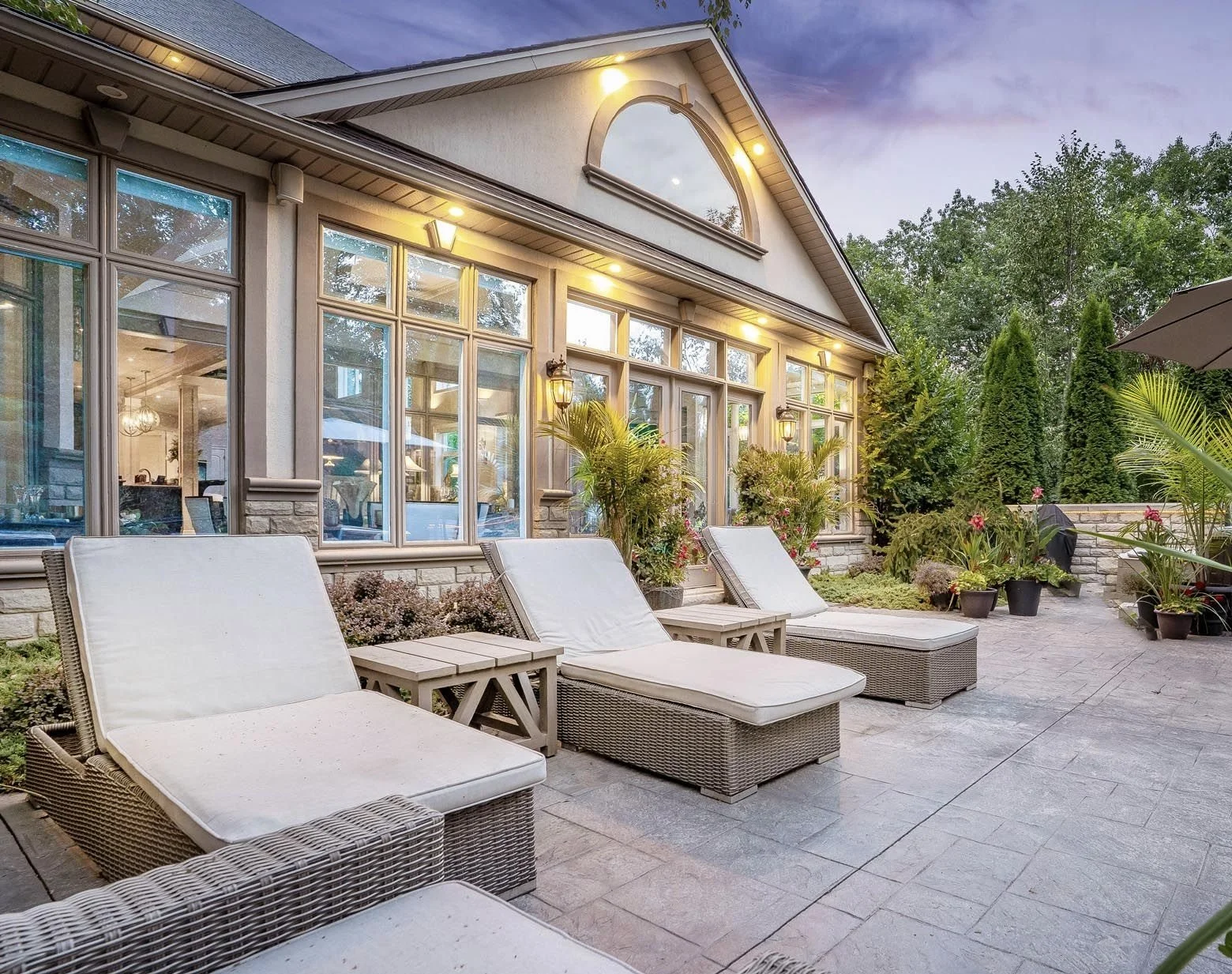Back patio with lounge chairs, potted plants, and outdoor lighting, in front of a large windowed house with trees in background, during dusk.