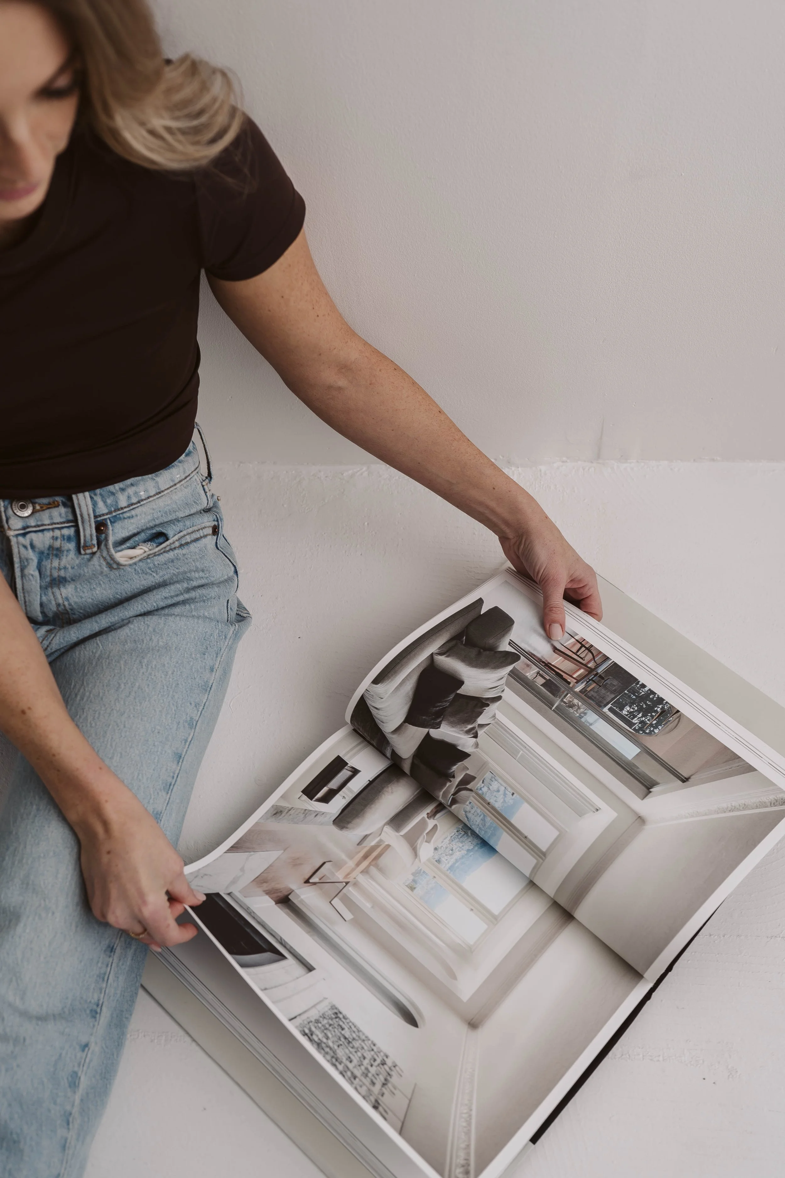 A woman with blond hair wearing a brown t-shirt and light blue jeans sits on the floor, flipping through a large home interior design magazine with images of a bright, modern living room decorating.