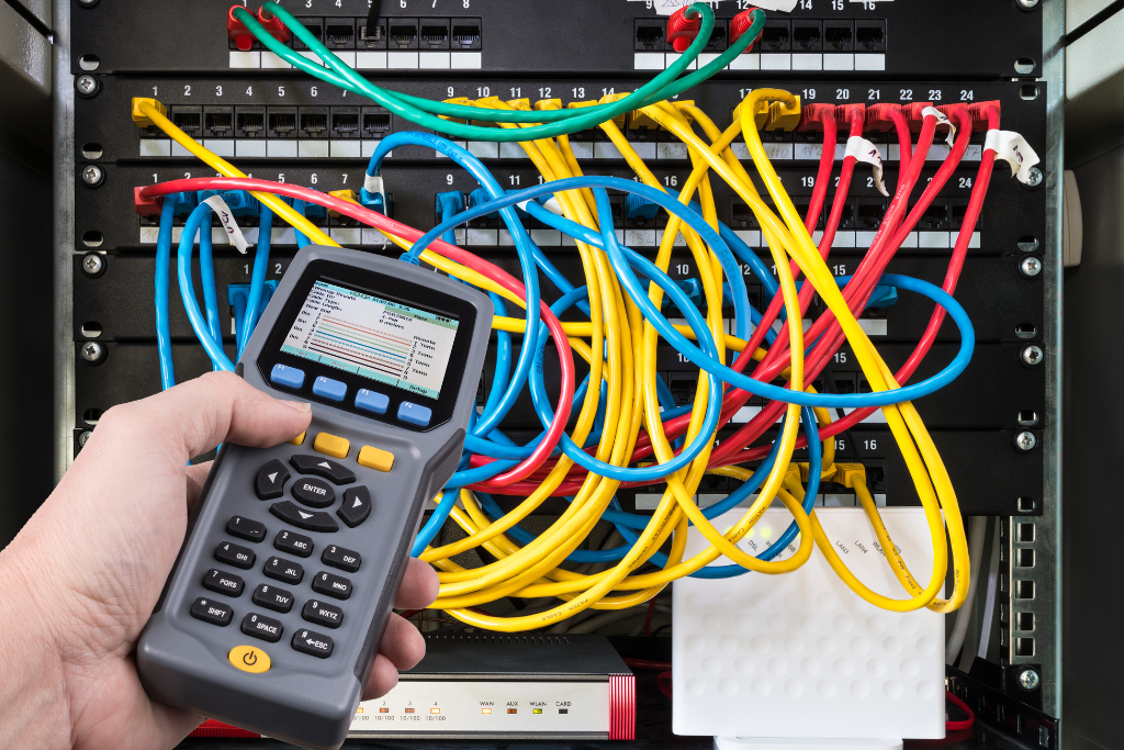 A person's hand holding a network testing device in front of a server rack with colorful Ethernet cables connected to switches.