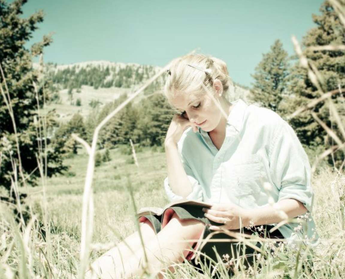 A woman sitting outdoors reading a book in a grassy field with trees and mountains in the background.