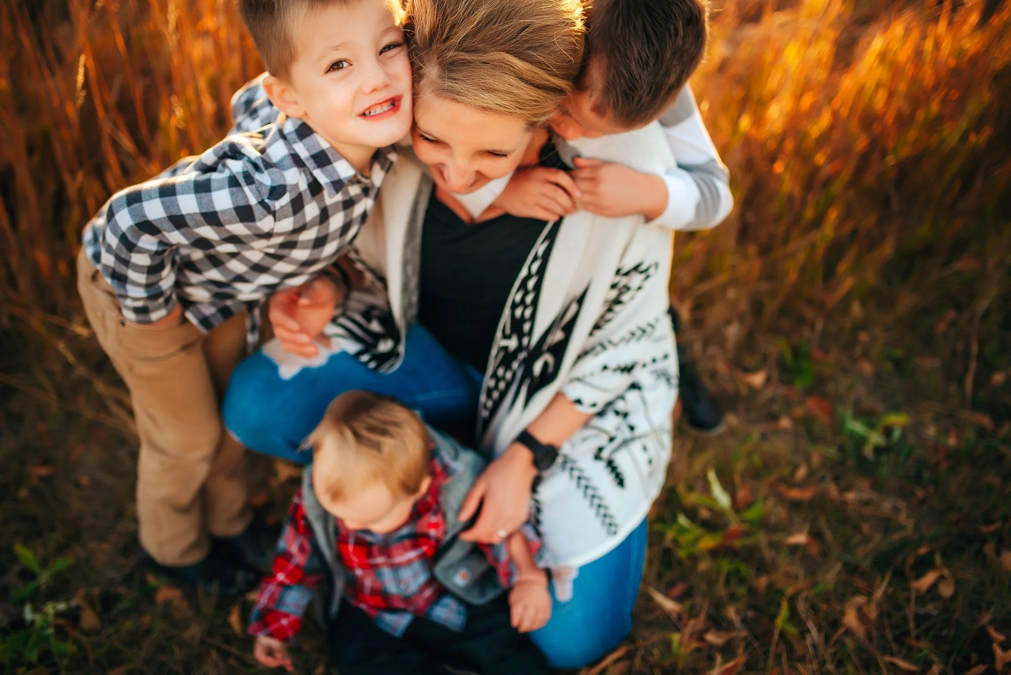 A woman and three children smiling and laughing outdoors in a grassy field with fall foliage, captured from above.