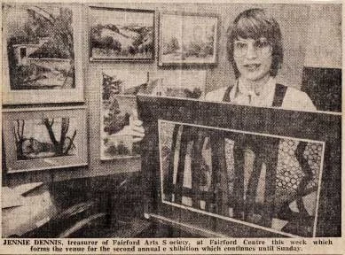 A woman standing behind a framed artwork, with several landscape paintings hanging on the wall behind her in an art gallery or store.