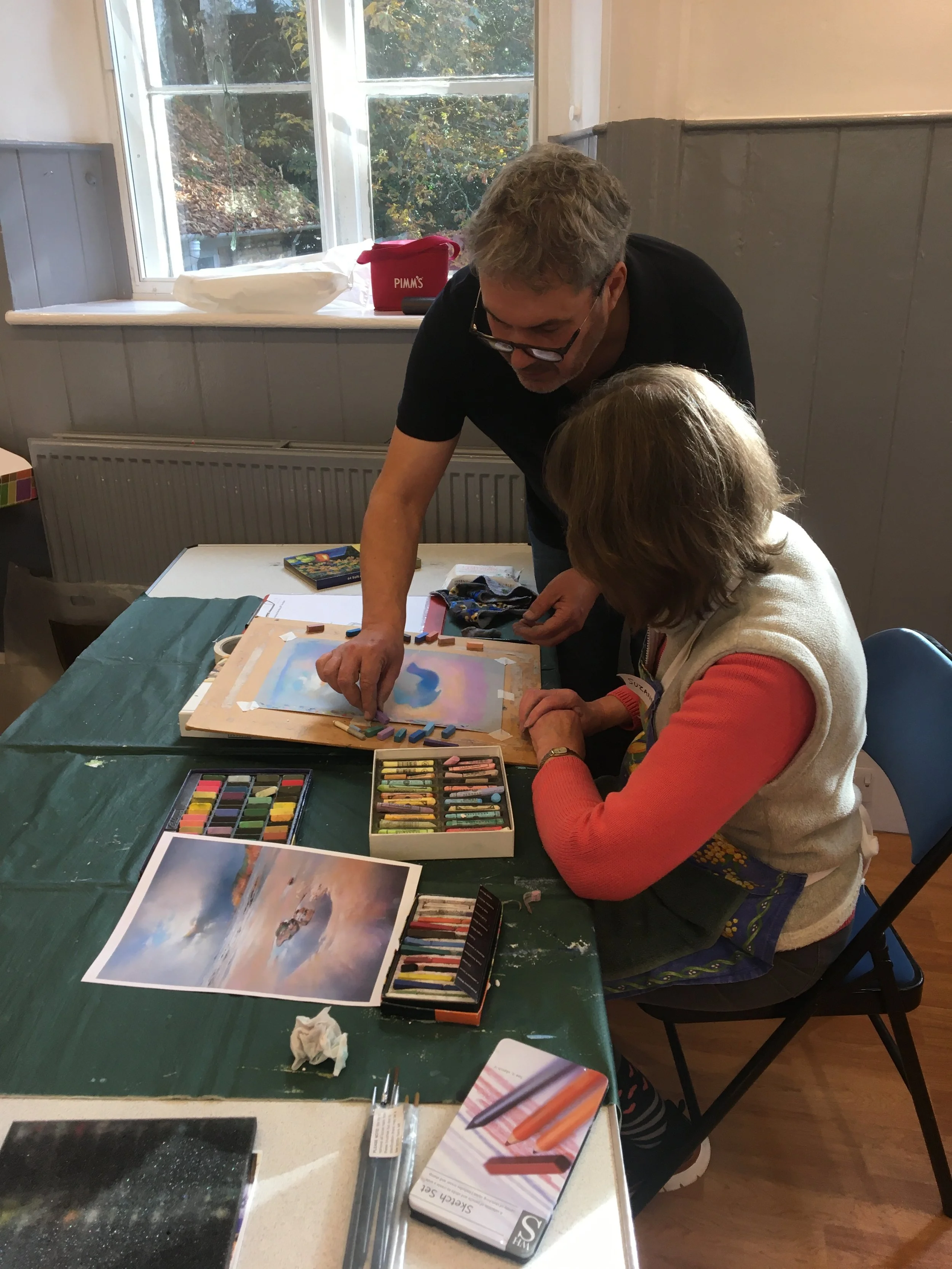A man and a woman are sitting at a table engaged in an art activity. The man is standing and leaning over, pointing at a piece of artwork, while the woman is seated, watching or listening. The table has various art supplies, including pastels, a picture of a floating island, and reference images. There is a window behind them showing greenery outside.