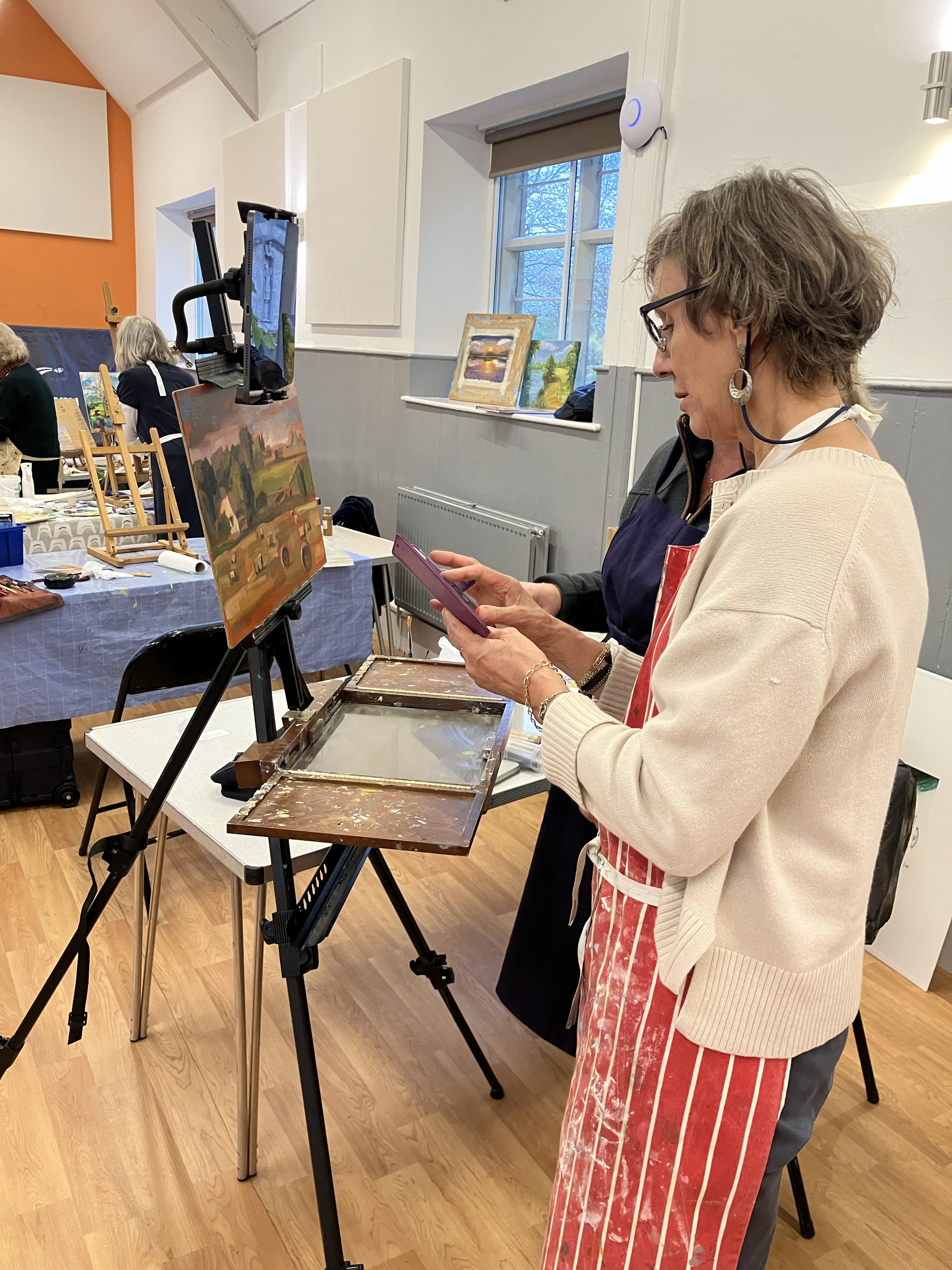 Woman in glasses and a striped apron looking at her phone while standing at an art class or workshop with paintings and art supplies around.