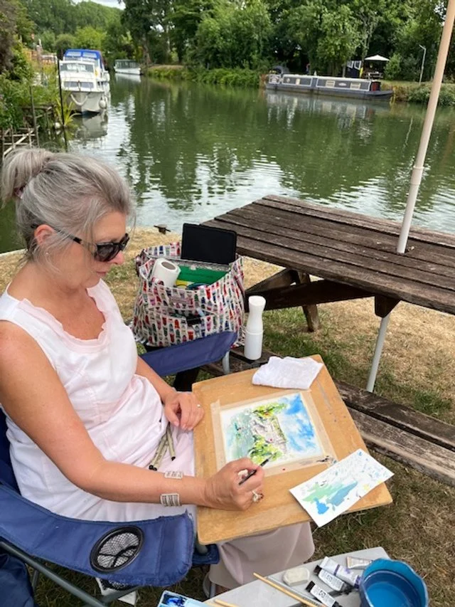 Woman sitting in a chair outdoors by a river, painting on a portable easel with boats and trees in the background.