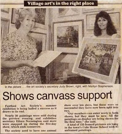 A black and white newspaper photo showing Judy Brown, the art society's secretary, and Marilyn Stephenson at an art exhibition. Judy Brown is on the left, holding an open book, with framed landscape paintings on display around them.