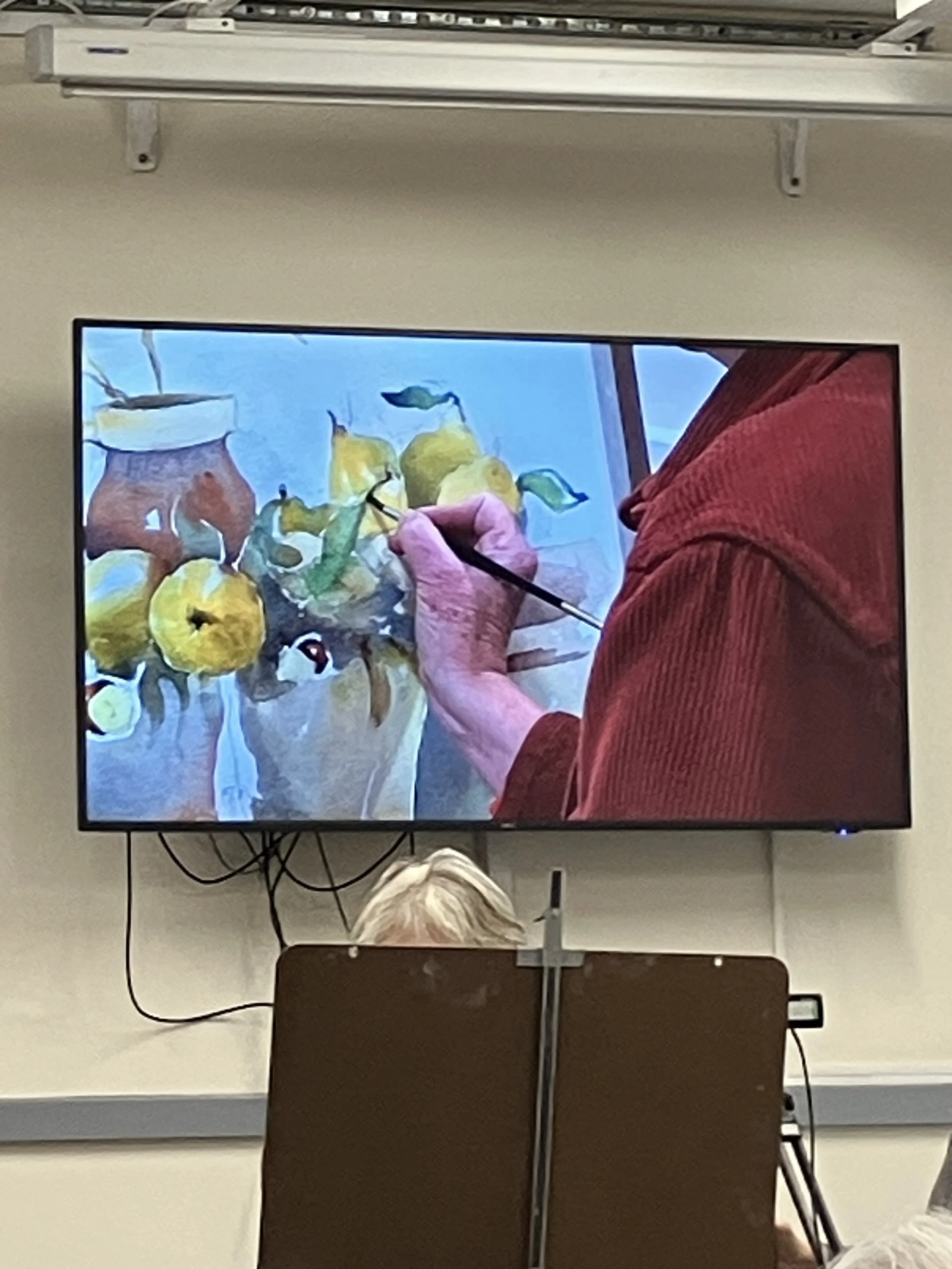 Person in a red shirt painting a still life with yellow pears and a pitcher on a computer monitor.
