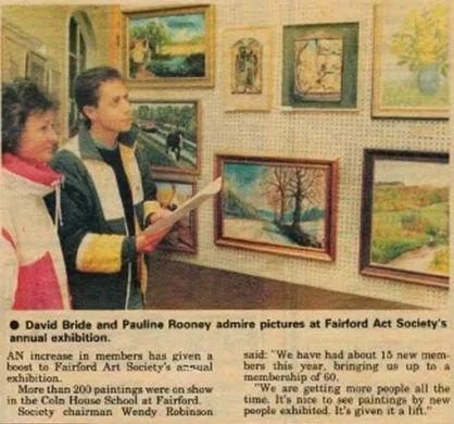 Two women standing in front of a wall displaying various landscape paintings, with one holding a brochure or program.