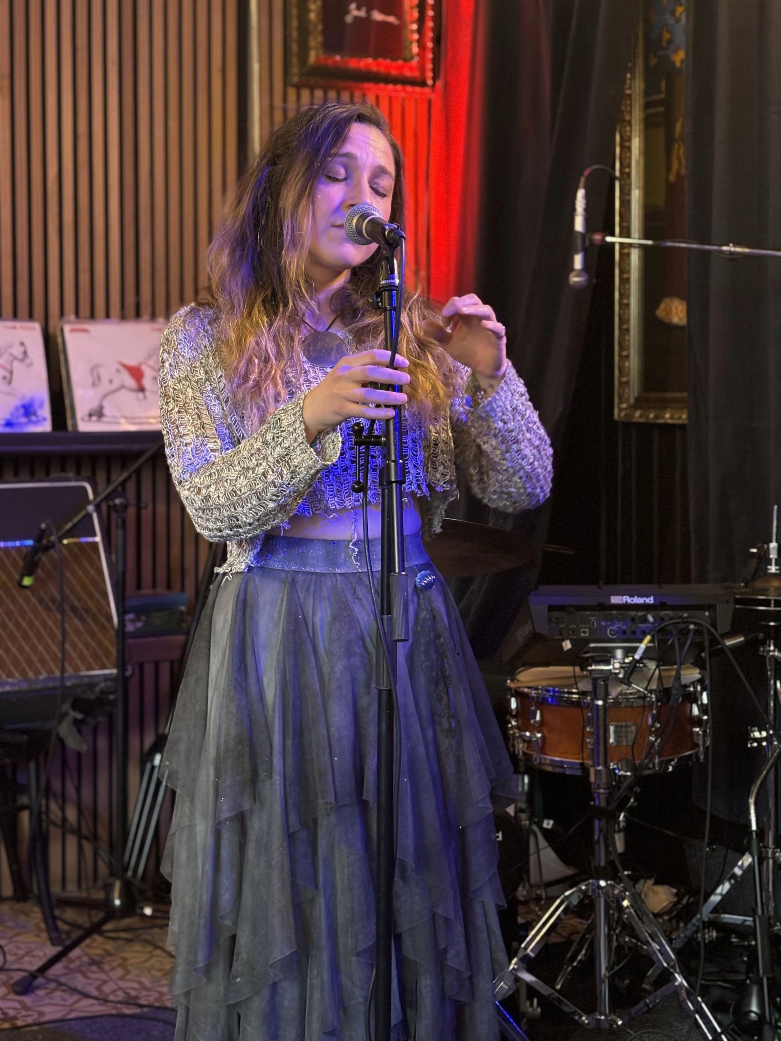 A woman with long hair singing or speaking into a microphone in a music venue or studio, with musical instruments and equipment in the background.