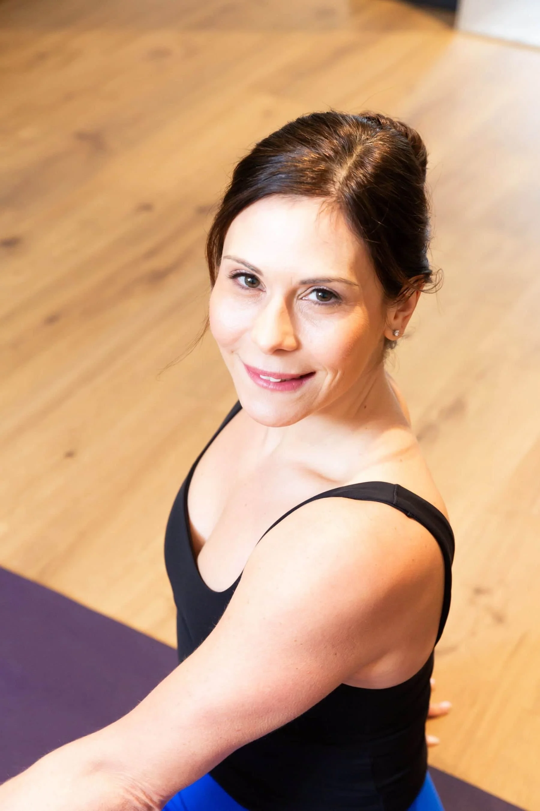 Woman with brown hair in a bun smiling and looking up at the camera, wearing a black tank top, sitting on a yoga mat on a wooden floor.