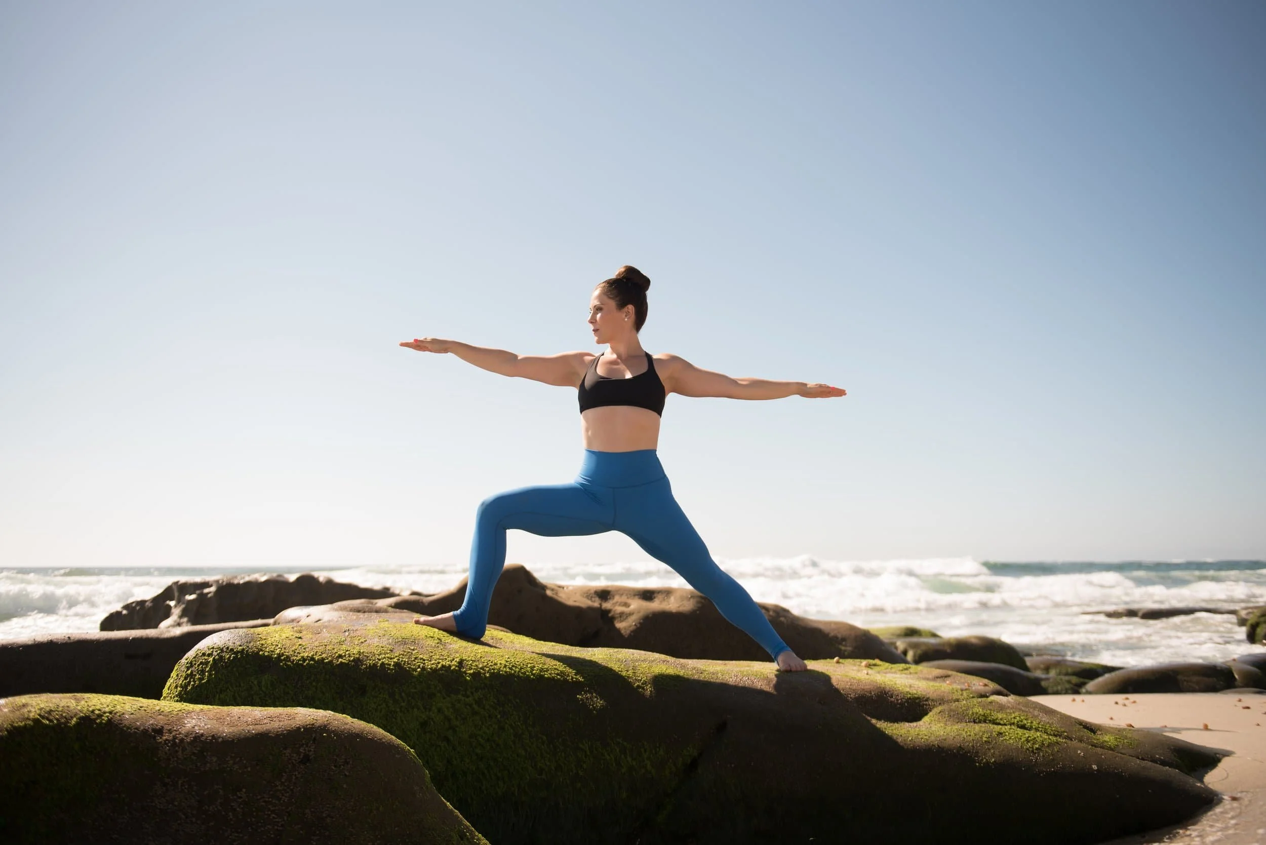 A woman practicing yoga outdoors on rocks by the beach during daylight, performing Warrior pose with arms extended and one foot forward.