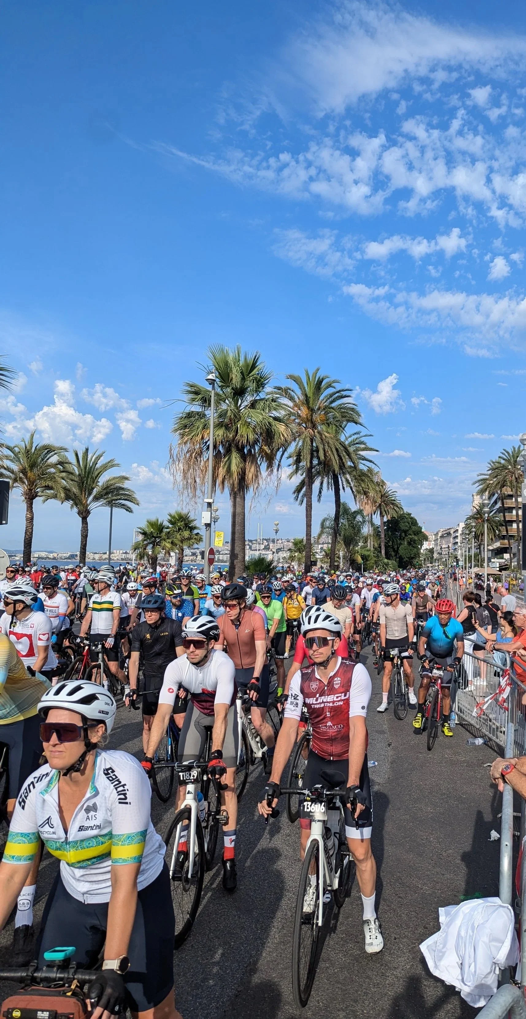 A large group of cyclists participating in a race or event near a coastal area, with palm trees, a clear blue sky, and a cityscape in the background.
