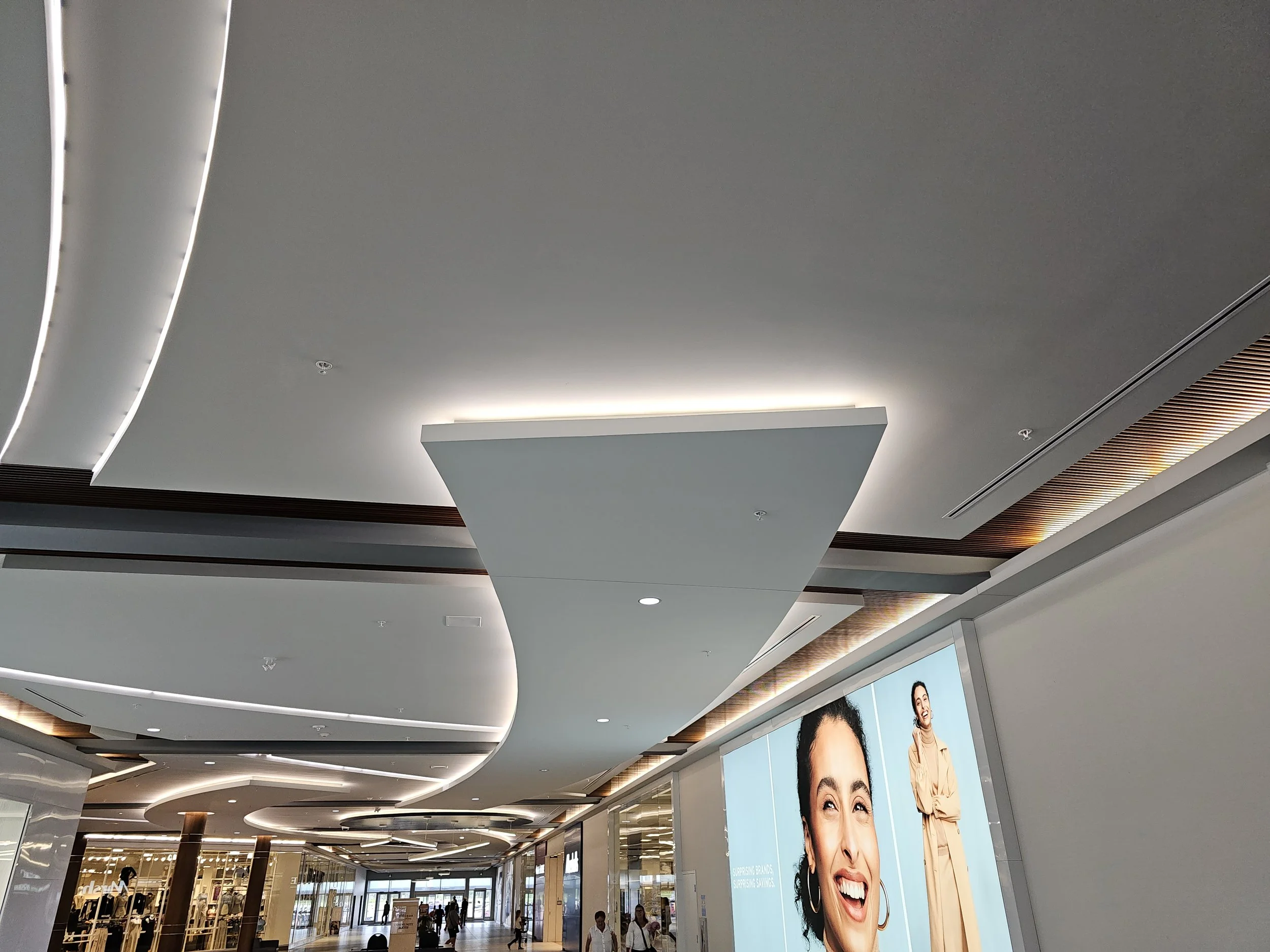 Interior of a modern shopping mall with curved ceiling designs, large digital advertisements, and shoppers walking in the corridor.