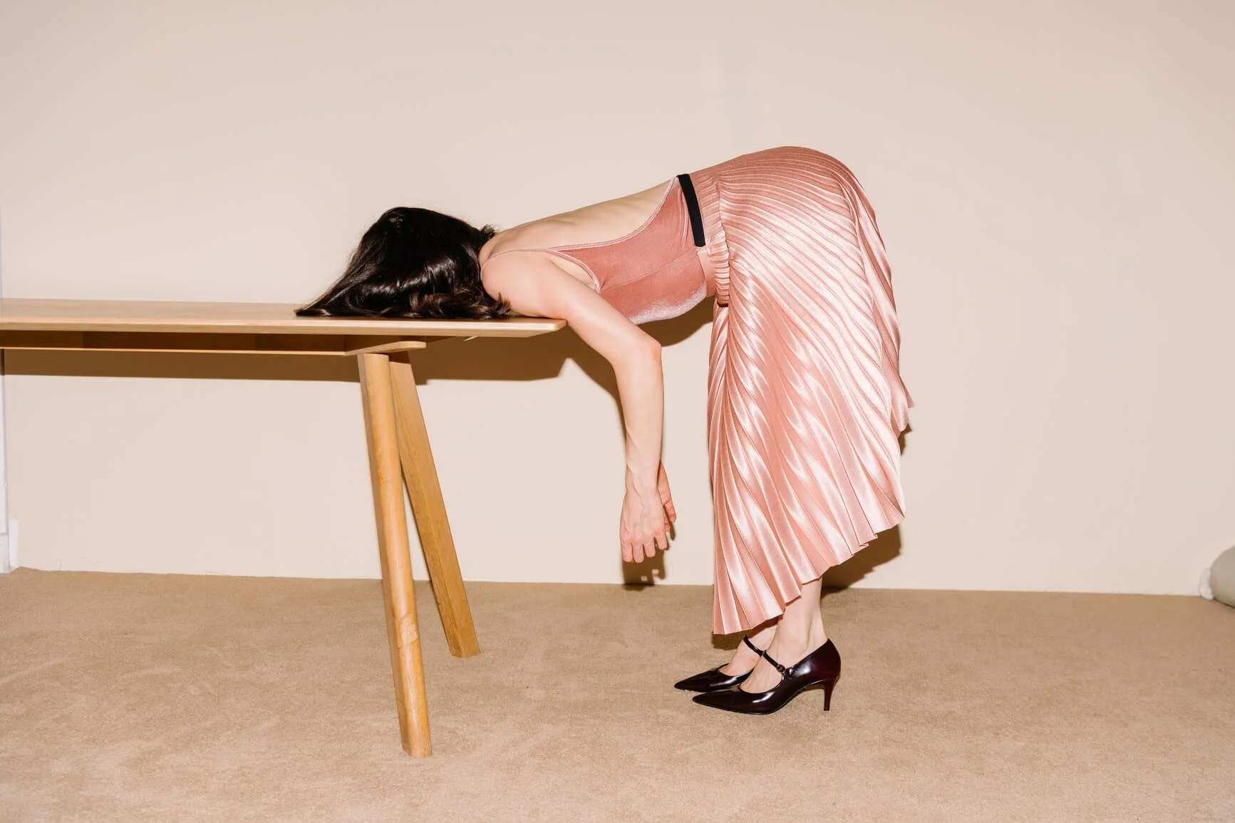 A person in a pleated pink dress and heels bends forward with their head resting on a wooden table in a minimalist room.
