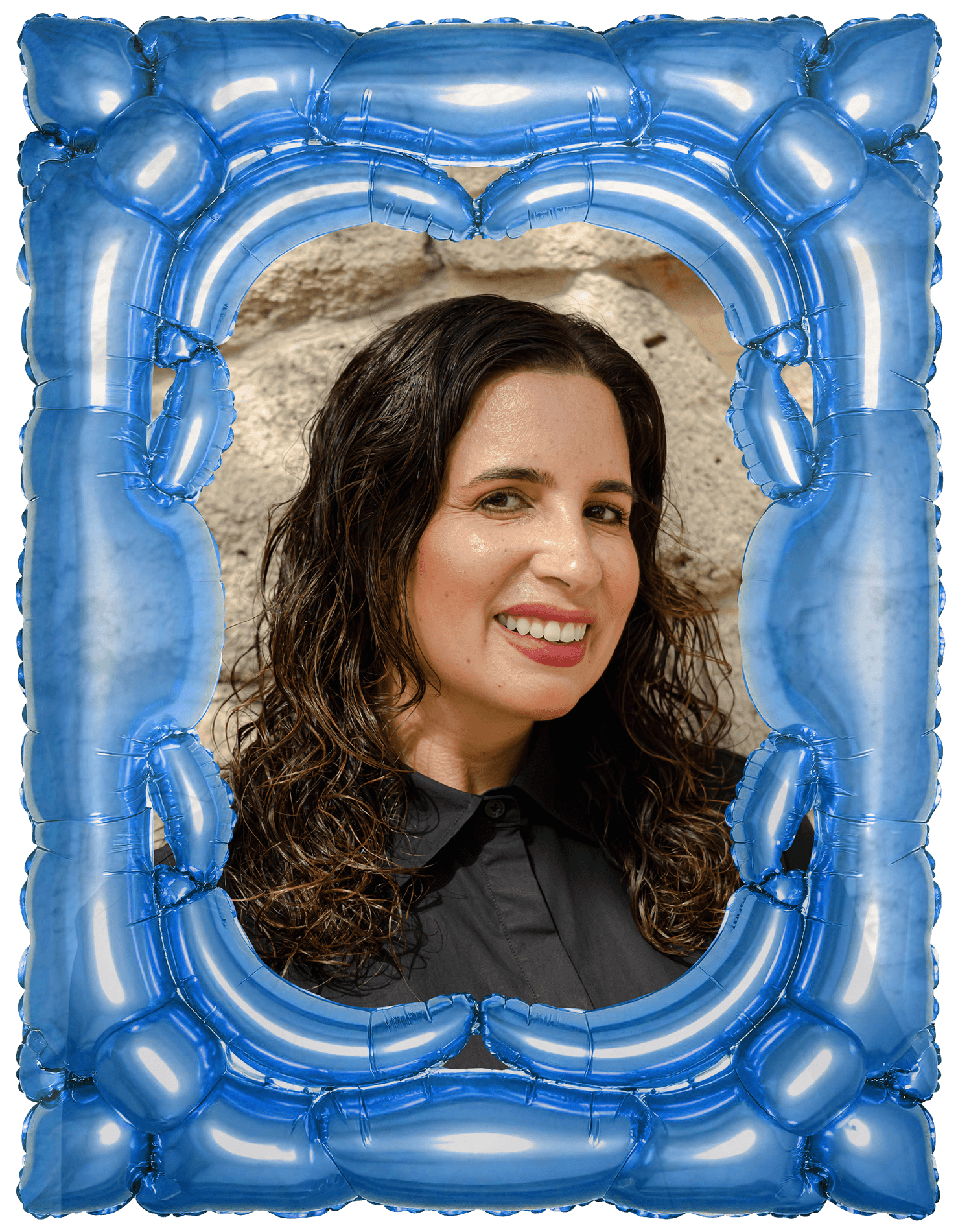 A woman with curly brown hair smiling, framed by a blue inflatable picture frame, standing in front of a stone wall.