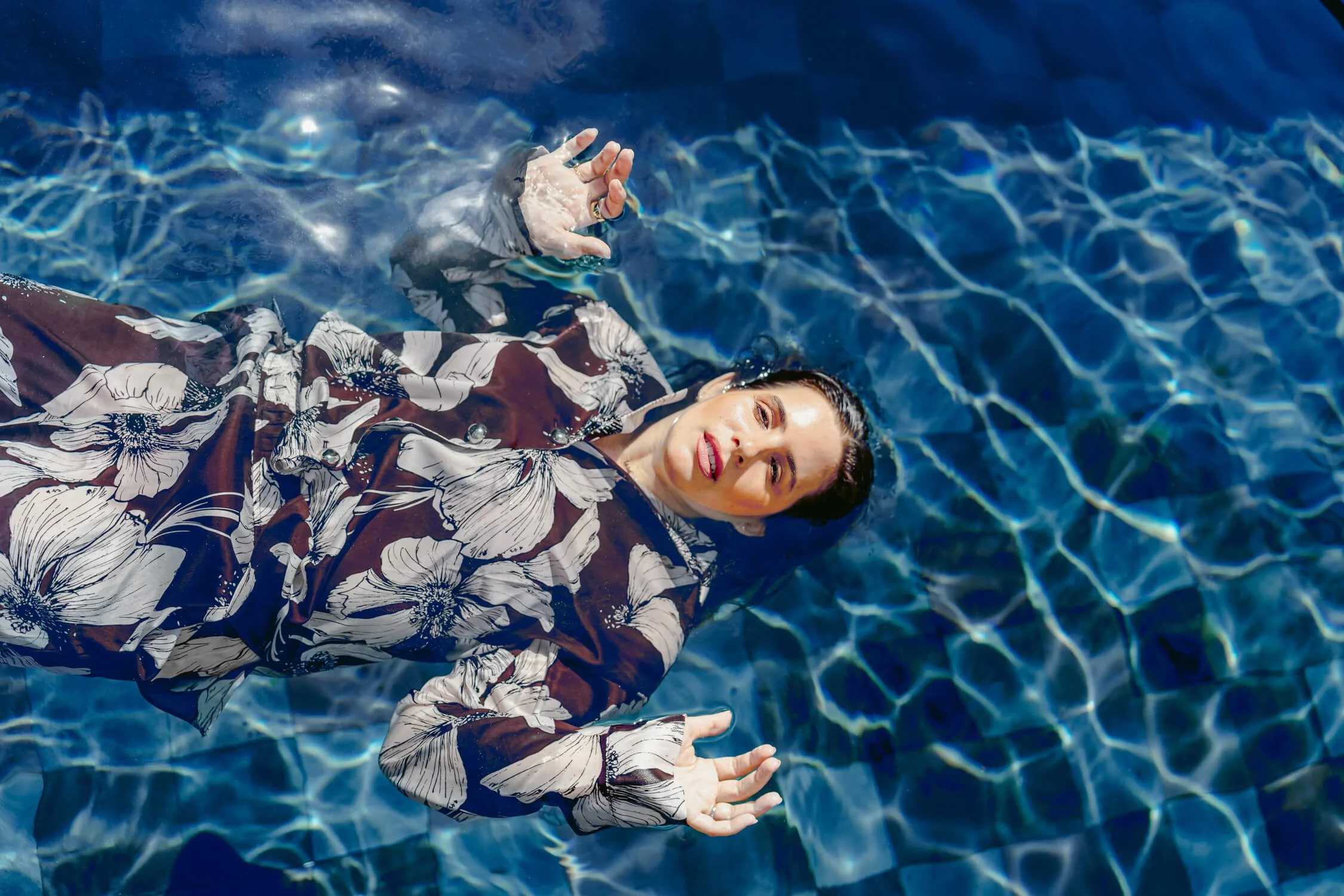 A woman  floats effortlessly in clear blue water, fully dressed, arms open, suspended between control and release.
