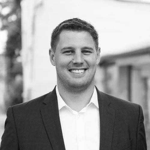 Black and white photo of a smiling man in a suit and white shirt, outdoors in front of a building.