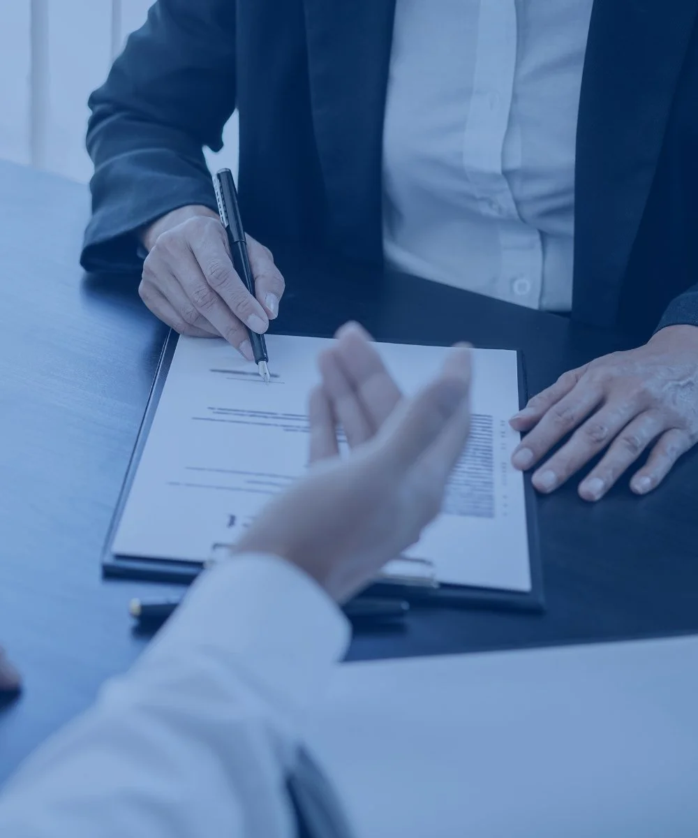 Two people in business attire are sitting at a table during a formal meeting. One person is signing a document with a black pen, while the other person discusses the document. The table has a dark surface and the setting appears professional.