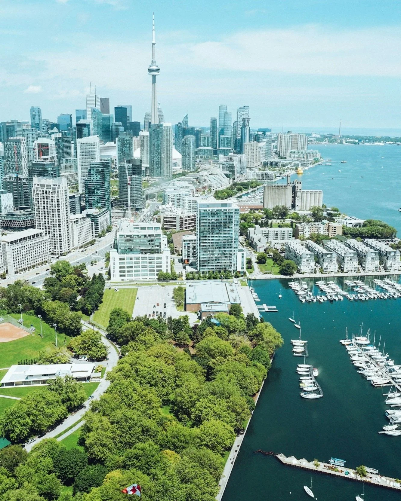 Aerial view of Toronto skyline with CN Tower, high-rise buildings, a park with trees, and a marina with boats along Lake Ontario.