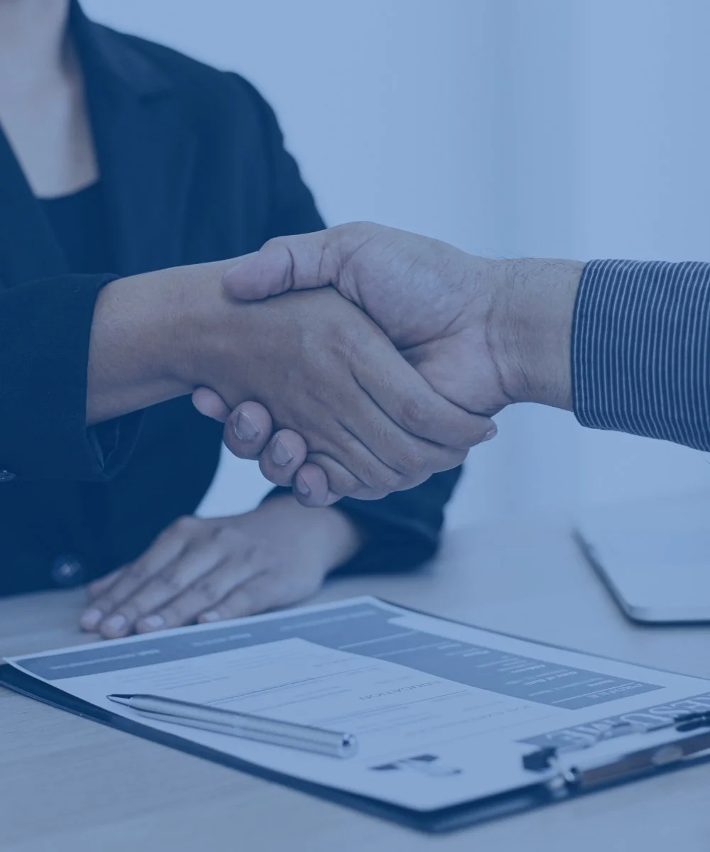 Two people, one with darker skin, shaking hands over a desk with documents and a pen, indicating a formal agreement or meeting.