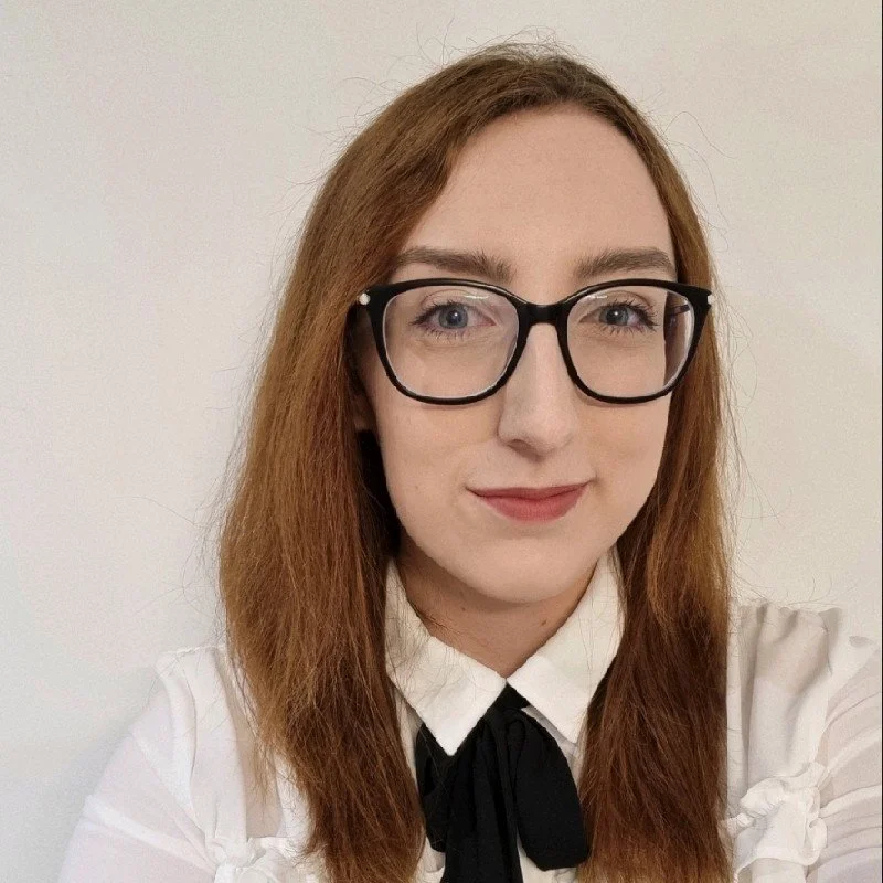 A young woman with light skin, long red hair, and wearing glasses with black frames. She is dressed in a white blouse with a black bow tie, standing in front of a plain white wall.