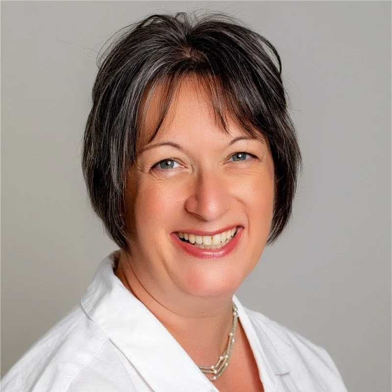 A smiling woman with short dark hair and blue eyes, wearing a white top and a pearl necklace, against a neutral background.