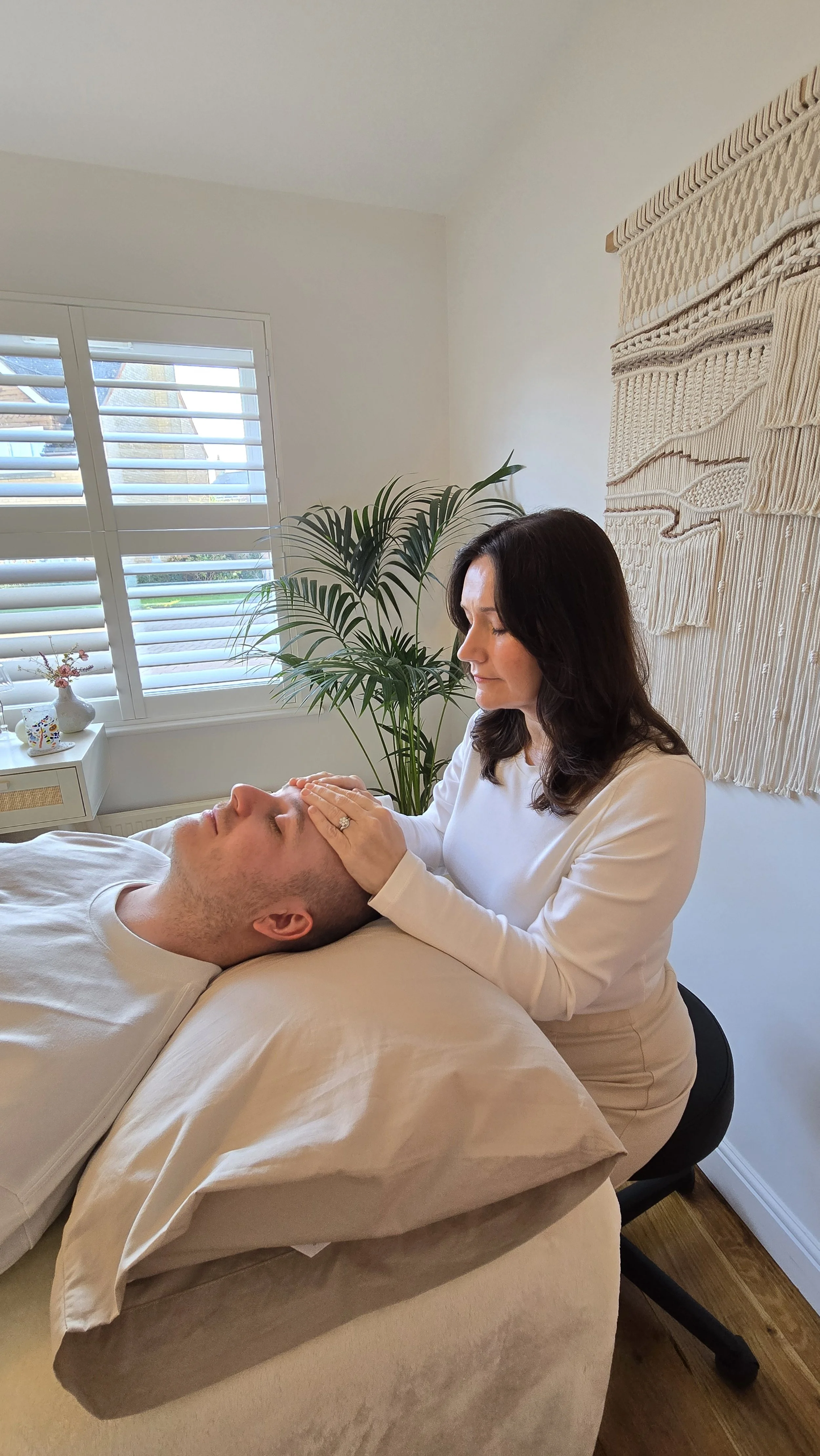 A woman providing a facial massage or treatment to a man lying on a massage table in a bright, cozy room with a large window, green plant, and decorative wall hanging.