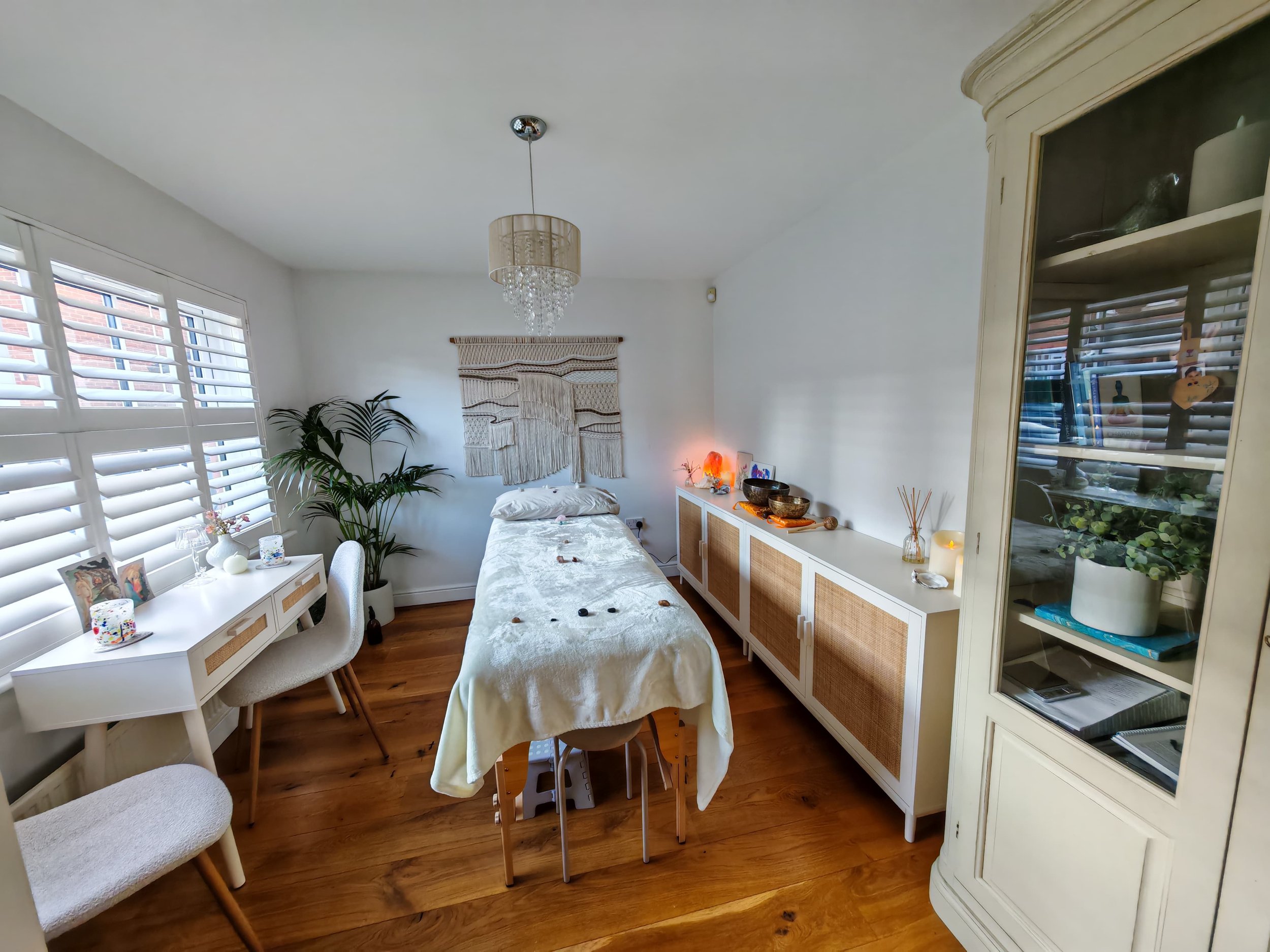 Divine Reiki's practice room with a massage table, surrounded by furniture, a large plant, and decorative items, with natural light coming through window blinds.