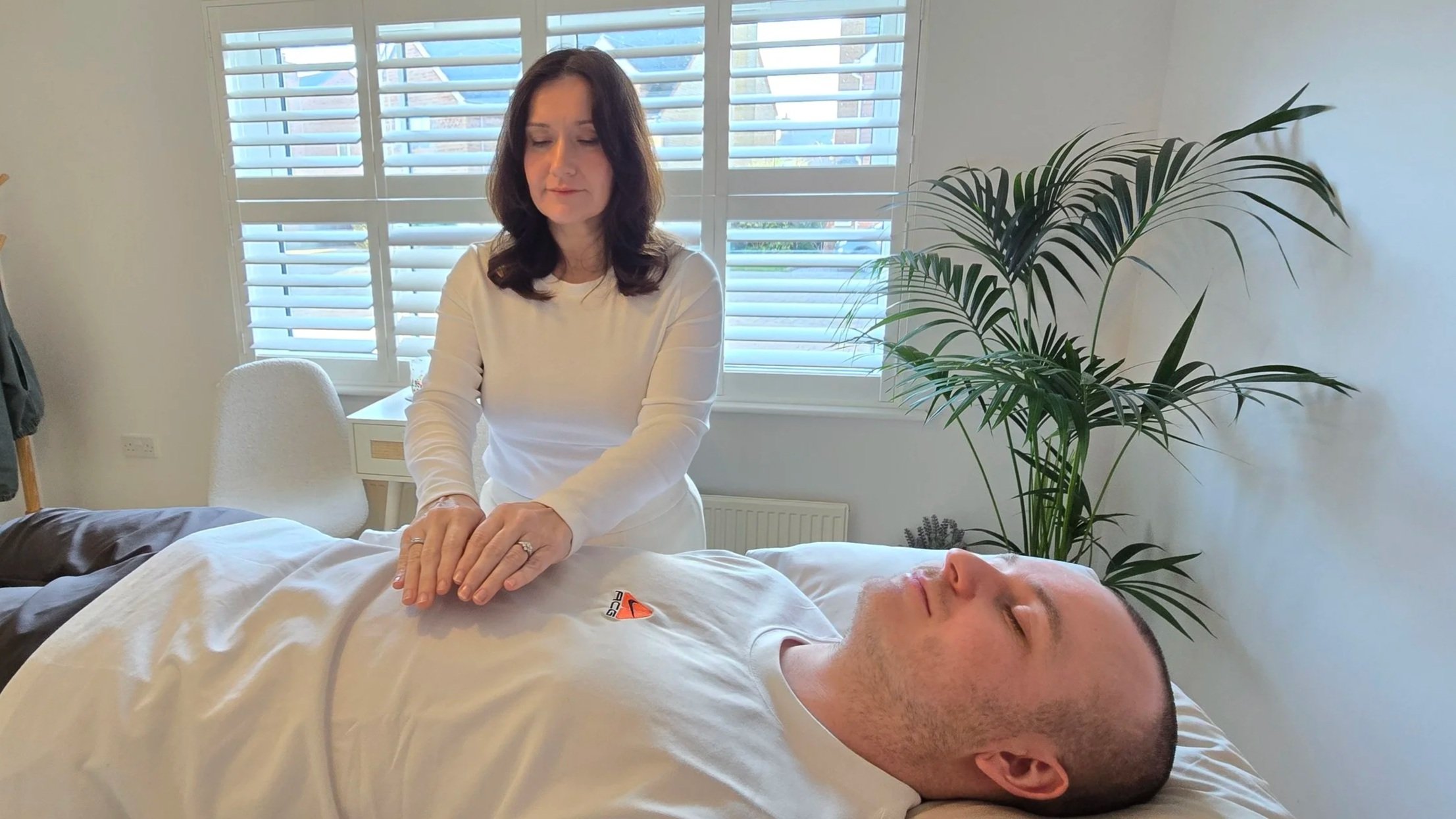 Jill from Divine Reiki is performing Reiki on a client lying on a bed in a well-lit room with large windows, blinds, and a green plant in the background.
