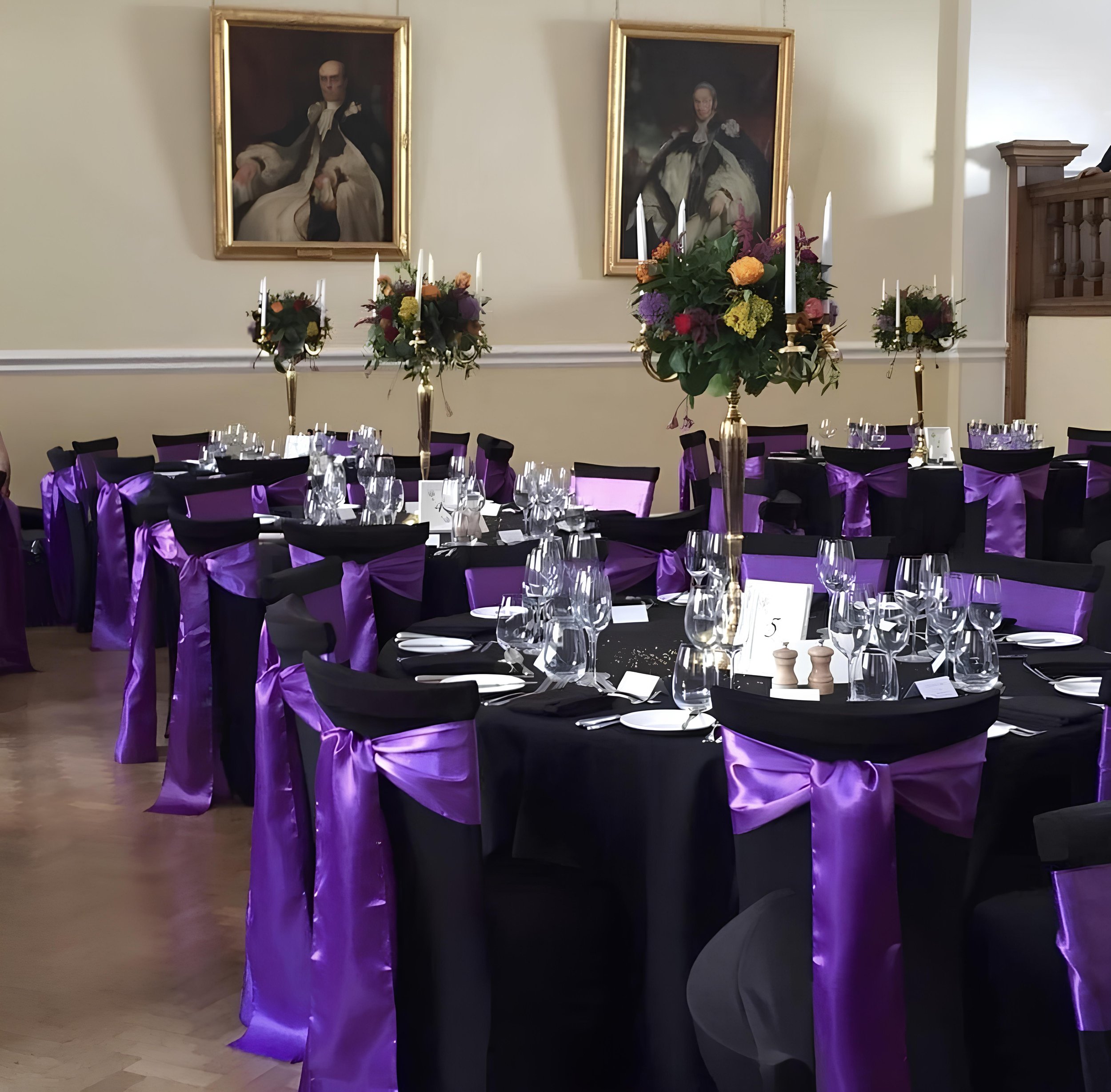 Elegant banquet hall decorated with round tables draped in black tablecloths, with tall gold vases holding floral arrangements as centerpieces. The chairs are adorned with black fabric covers and large purple satin bows. The tables are set with white plates, silverware, and multiple wine glasses. The background features two framed portraits of historical figures hanging on a cream-colored wall.
