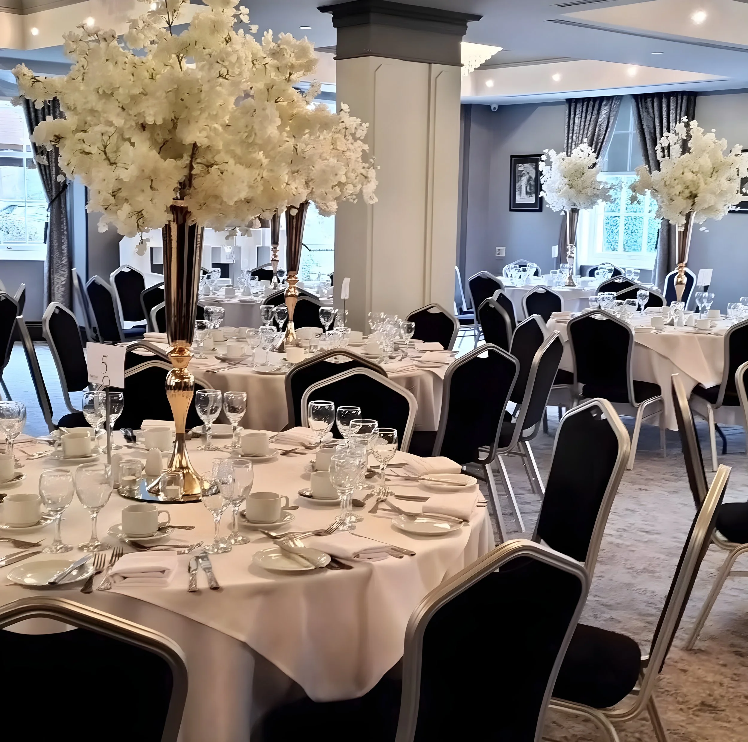 Elegant banquet hall decorated with white floral centerpieces on round tables with settings of glassware, china, and silverware, surrounded by black chairs with white trim.