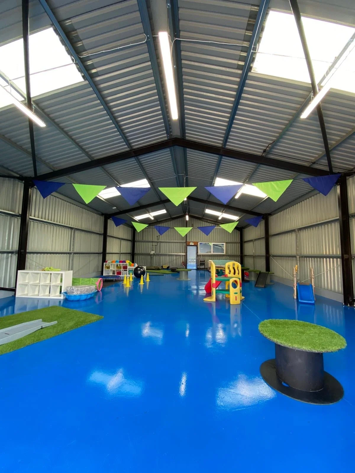 Indoor play area with blue flooring, colorful play structures, and decorations including blue and green pennant banners hanging from the ceiling.