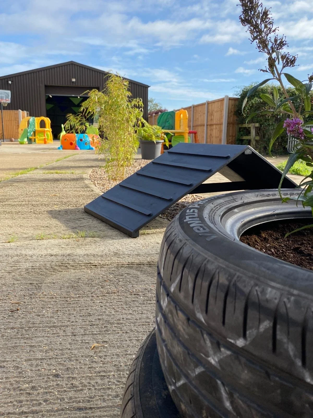 A backyard with a black wooden ramp leading to a planter with soil and a plant. In the background, there are colorful playground toys, a black shed, and a wooden fence under a blue sky with some clouds.
