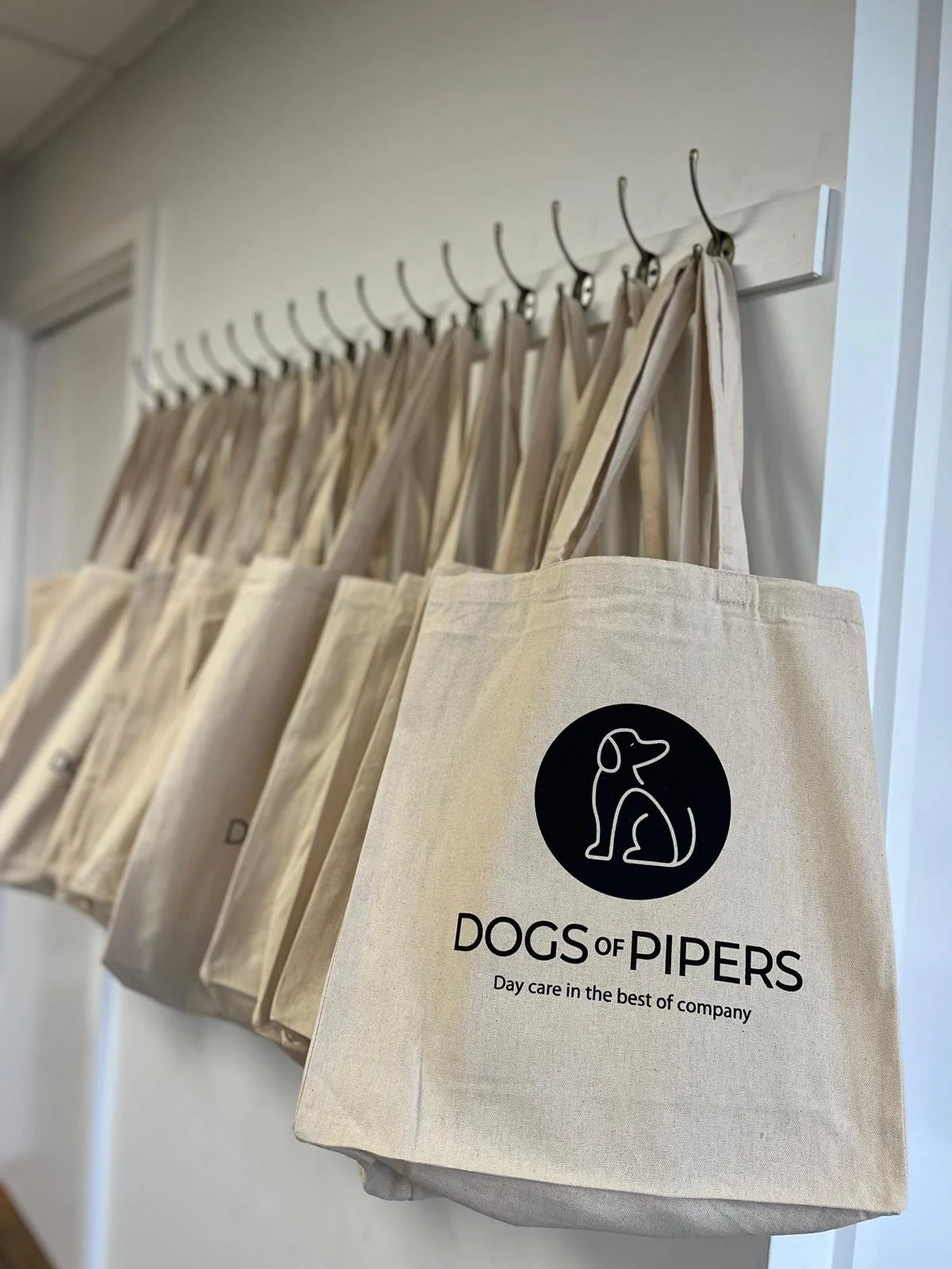 Beige tote bags hanging on hooks with the logo and text: "Dogs of Pipers - Day care in the best of company".