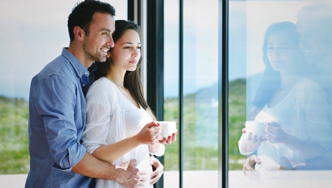 Un homme et une femme regardant par une grande fenêtre, tenant des tasses à café, dans un espace lumineux avec vue sur la campagne.