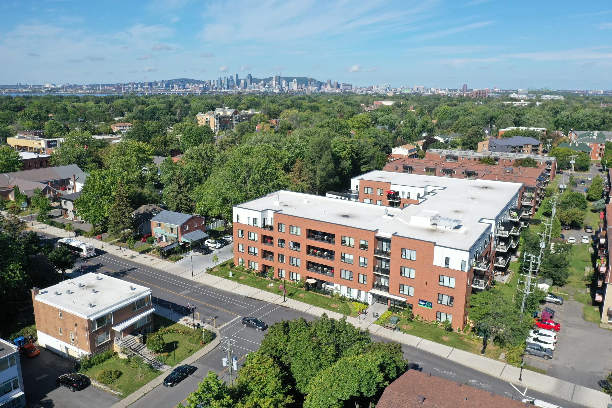 Vue aérienne d'un quartier résidentiel avec des immeubles modernes en briques rouges, des maisons et beaucoup d'arbres verts, avec la ville en arrière-plan sous un ciel bleu avec quelques nuages.