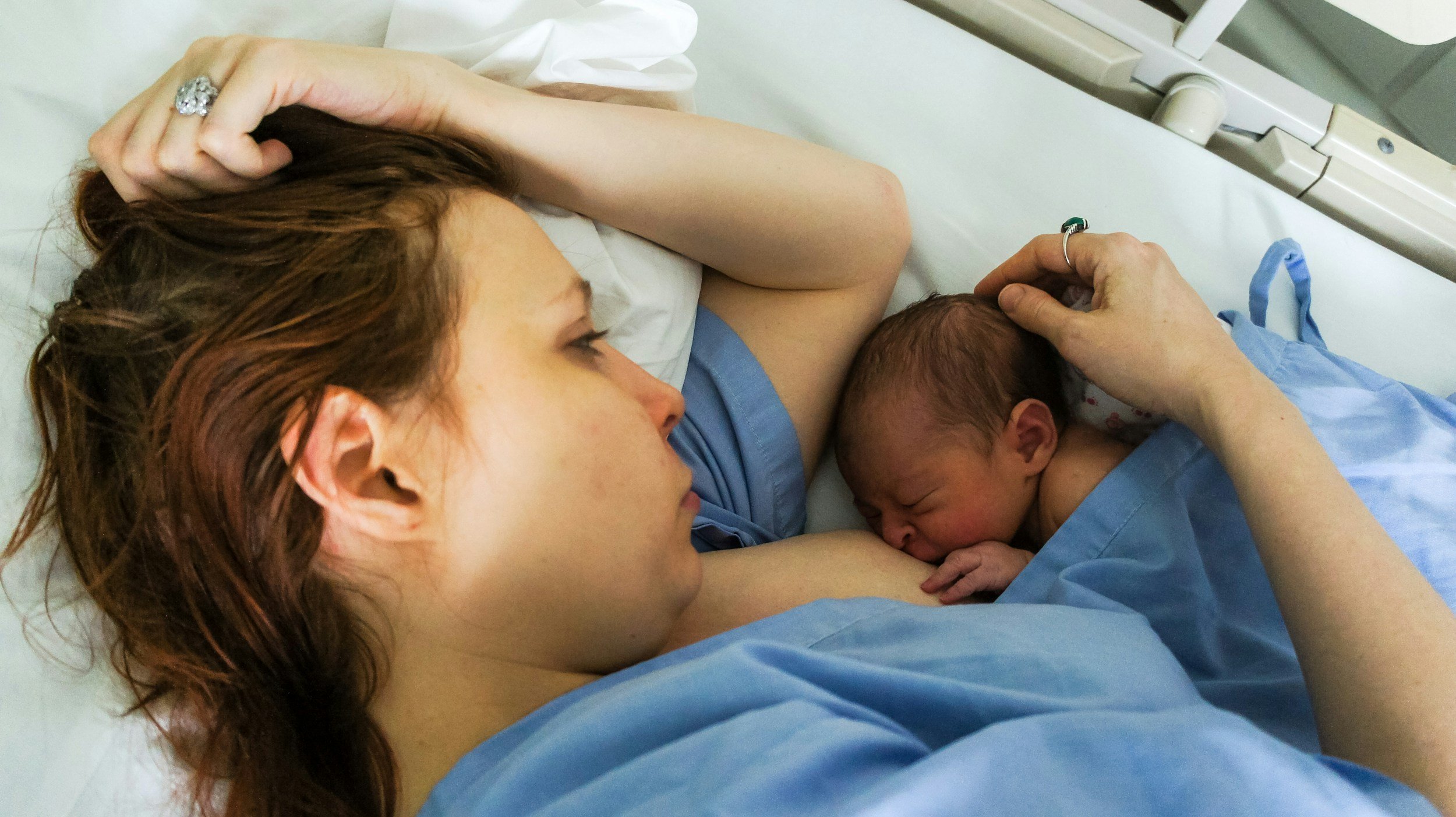 A new mother rests in a hospital bed, breastfeeding her newborn baby while holding the baby's head gently. Both are dressed in hospital gowns, and the scene suggests a peaceful moment after childbirth.