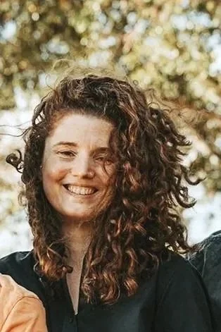 A woman with curly hair smiling outdoors with trees in the background.